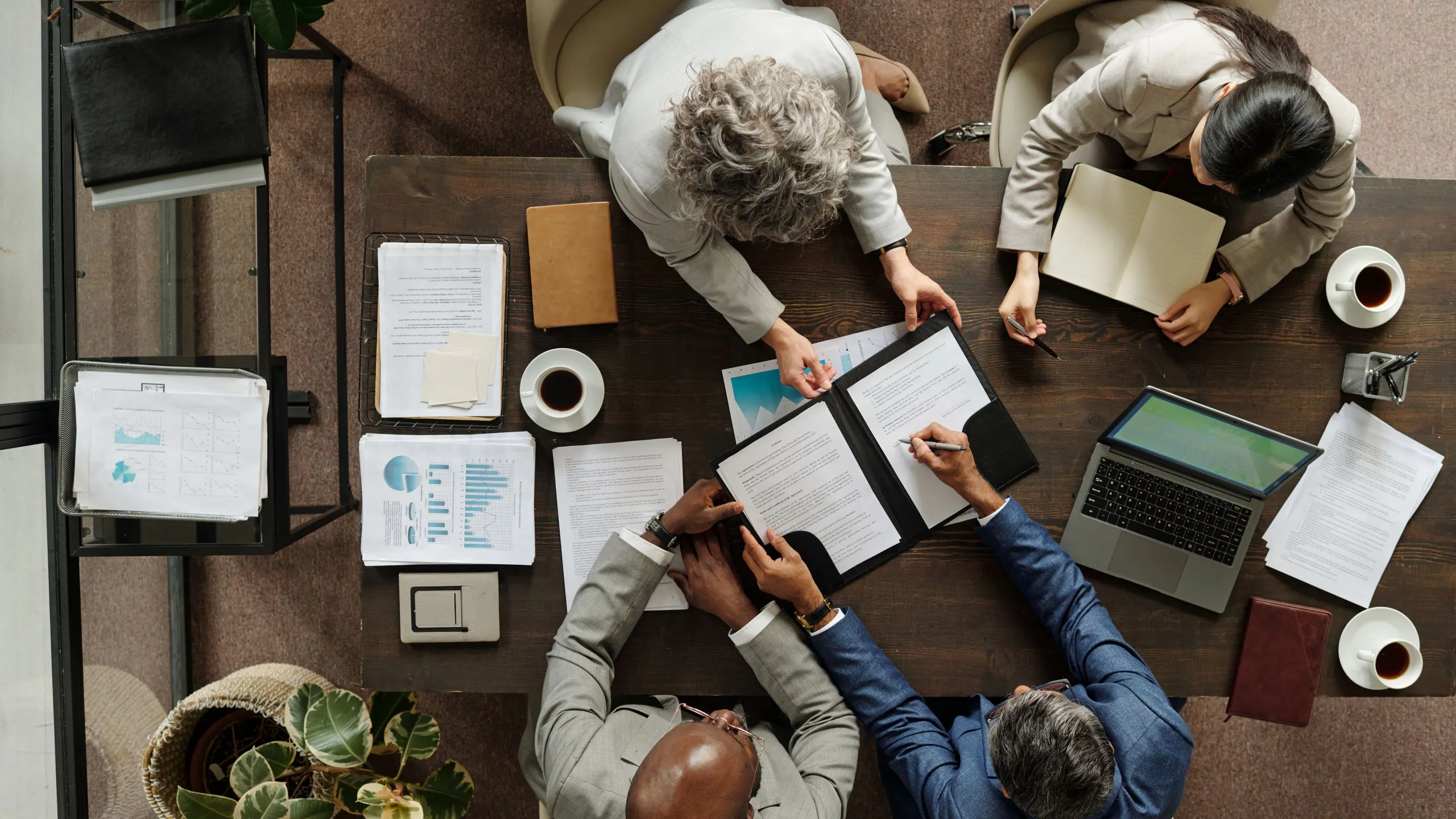 Overhead view of four professionals reviewing documents at a table with laptop, charts, and coffee during a meeting