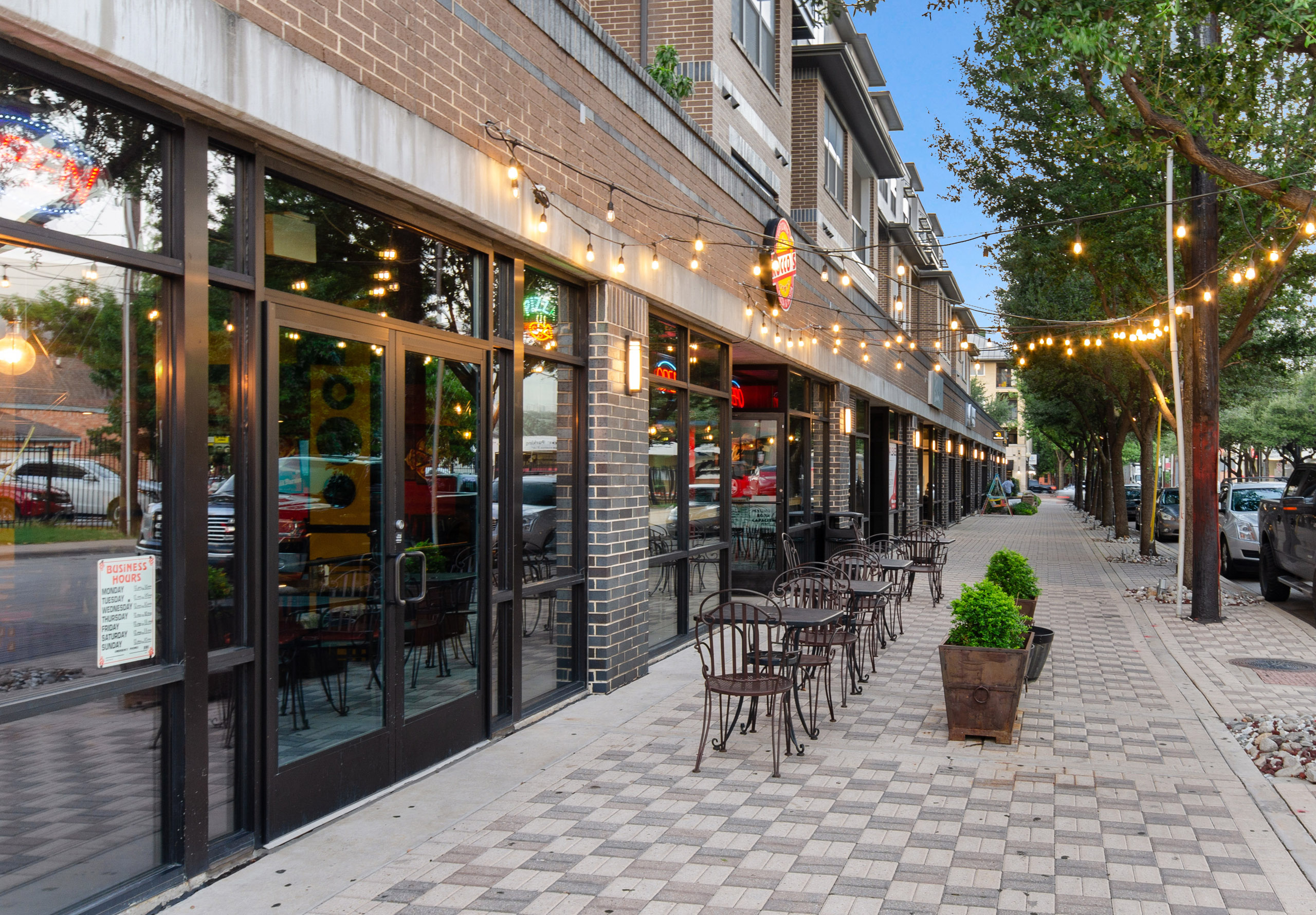 Exterior view at dusk of retailers at AMLI Quadrangle including a pizza restaurant with string lights and outdoor seating