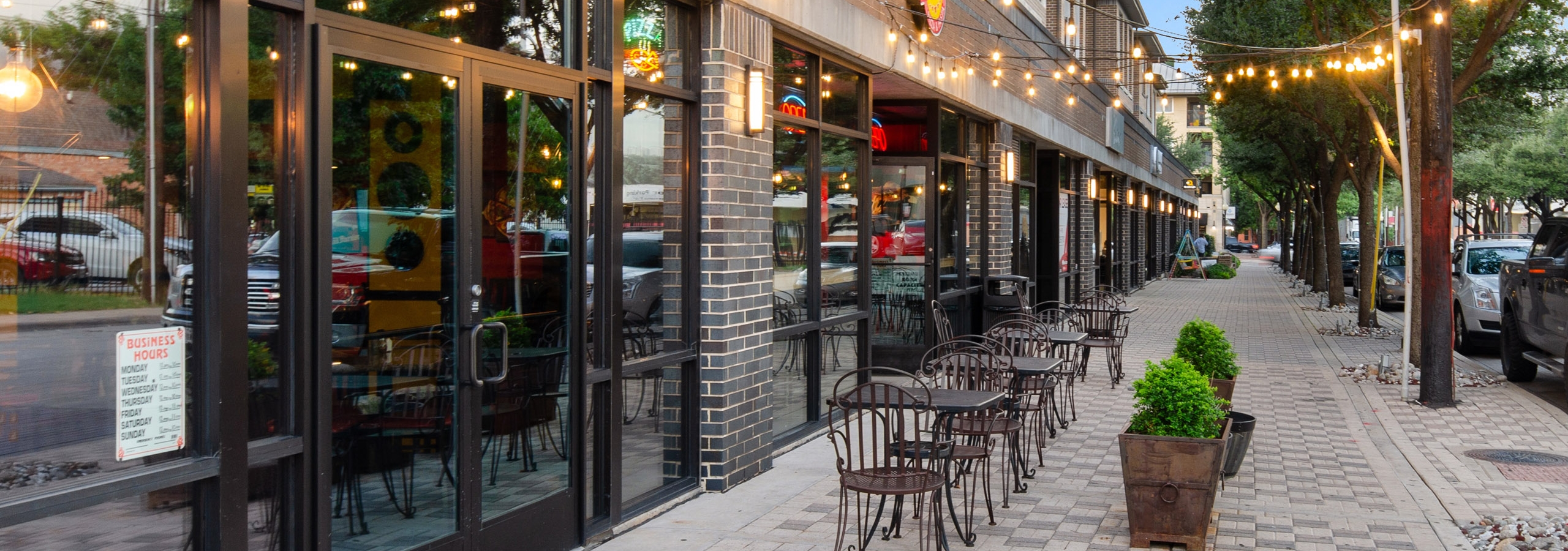 Exterior view at dusk of retailers at AMLI Quadrangle including a pizza restaurant with string lights and outdoor seating