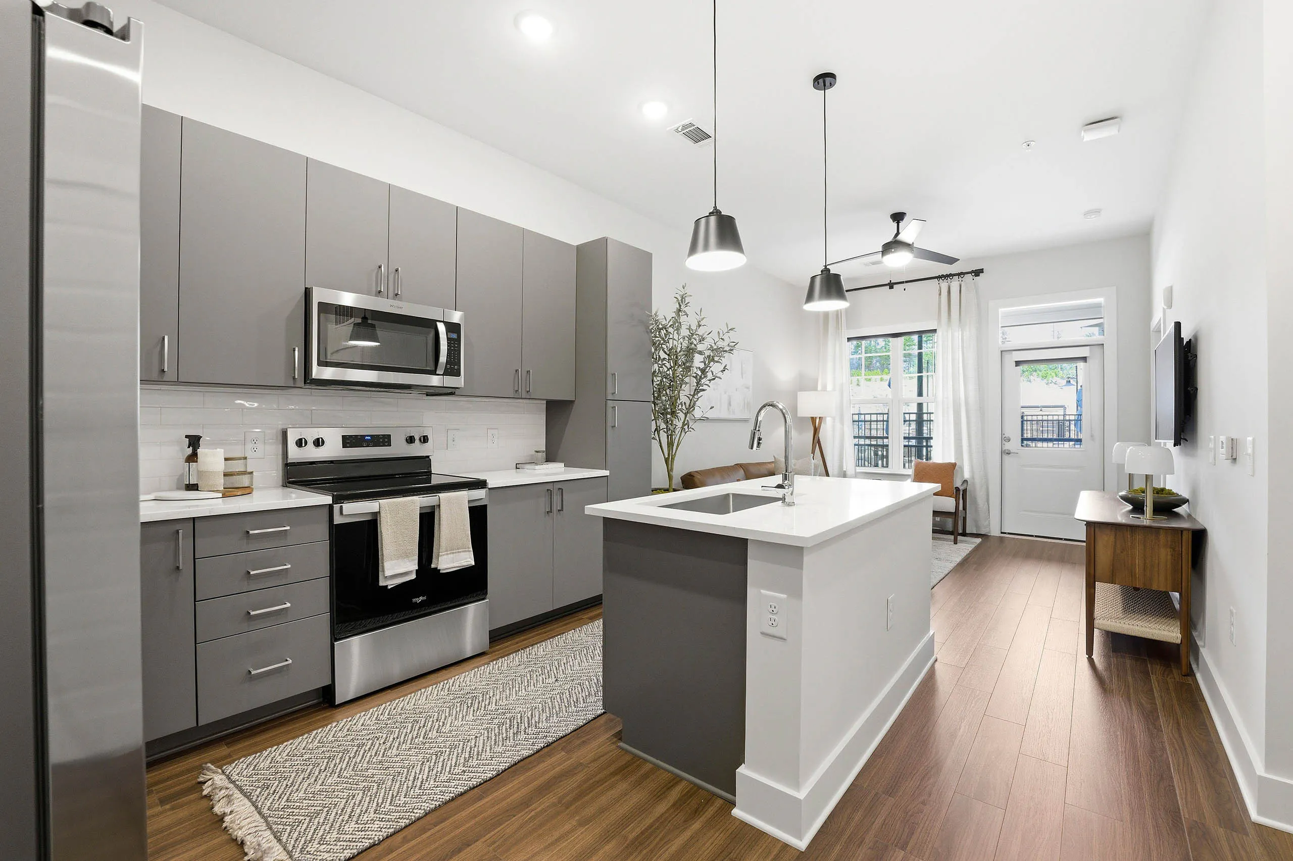 Modern kitchen with gray cabinets, stainless steel appliances, white island with sink, hanging lights overtop, open view to bright living area