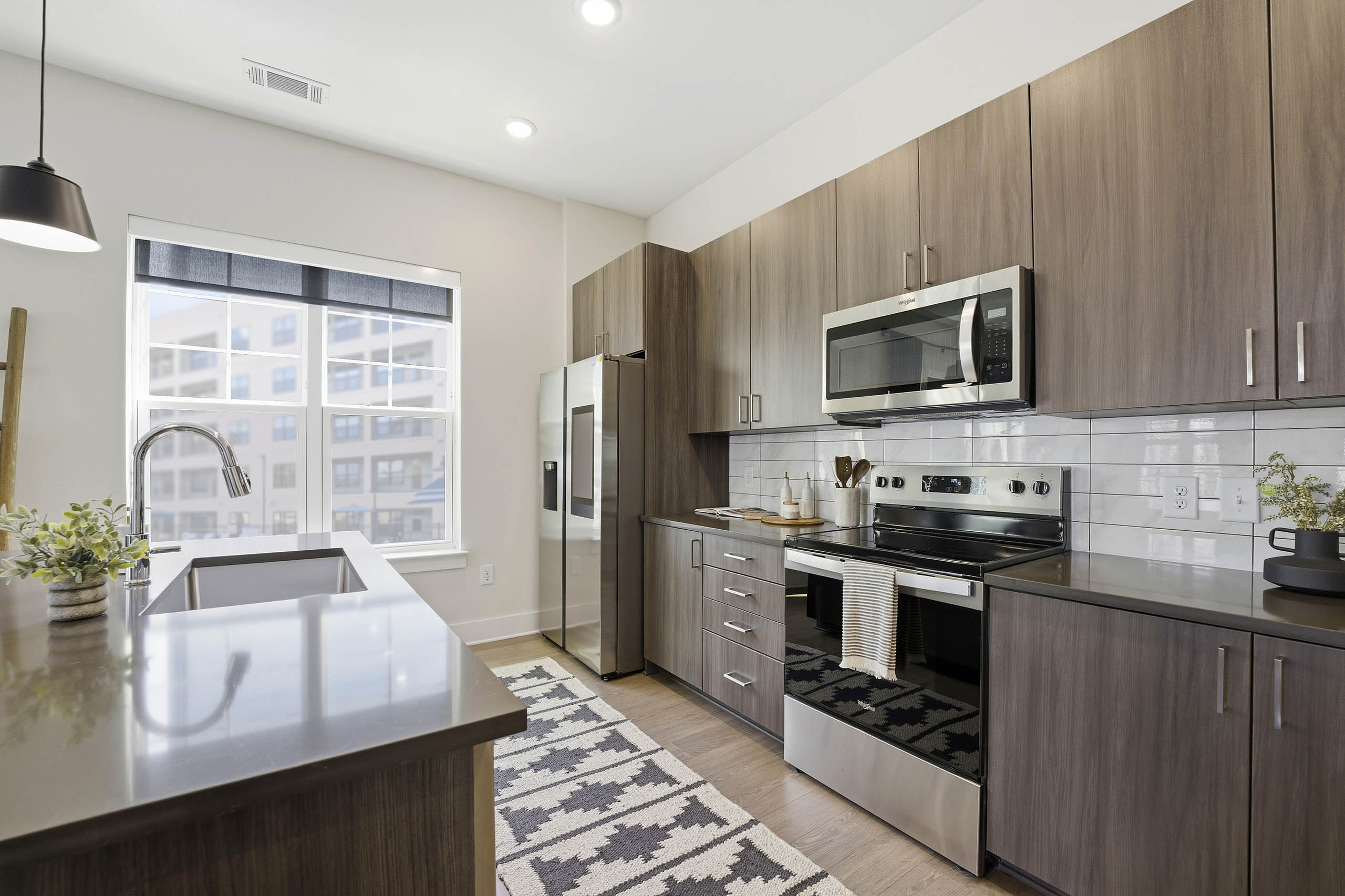 Kitchen with brown wood cabinetry, stainless steel appliances, brown island with sink, patterned cream and black rug, and large two pane window
