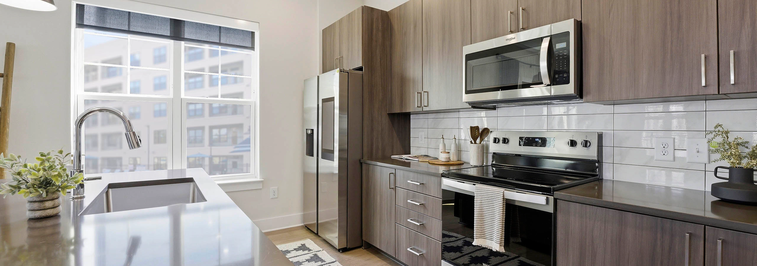 Kitchen with brown wood cabinetry, stainless steel appliances, brown island with sink, patterned cream and black rug, and large two pane window