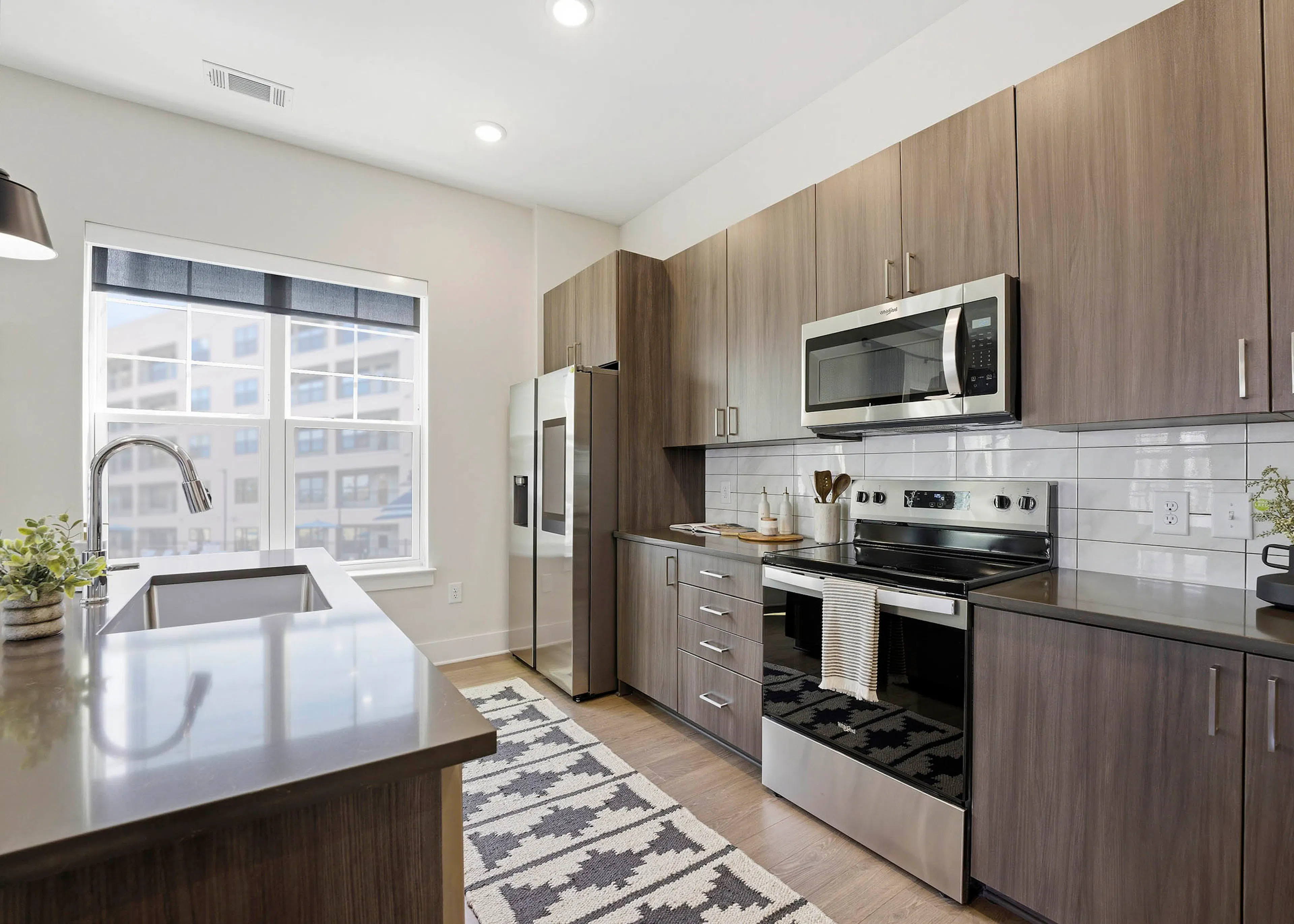 Kitchen with brown wood cabinetry, stainless steel appliances, brown island with sink, patterned cream and black rug, and large two pane window
