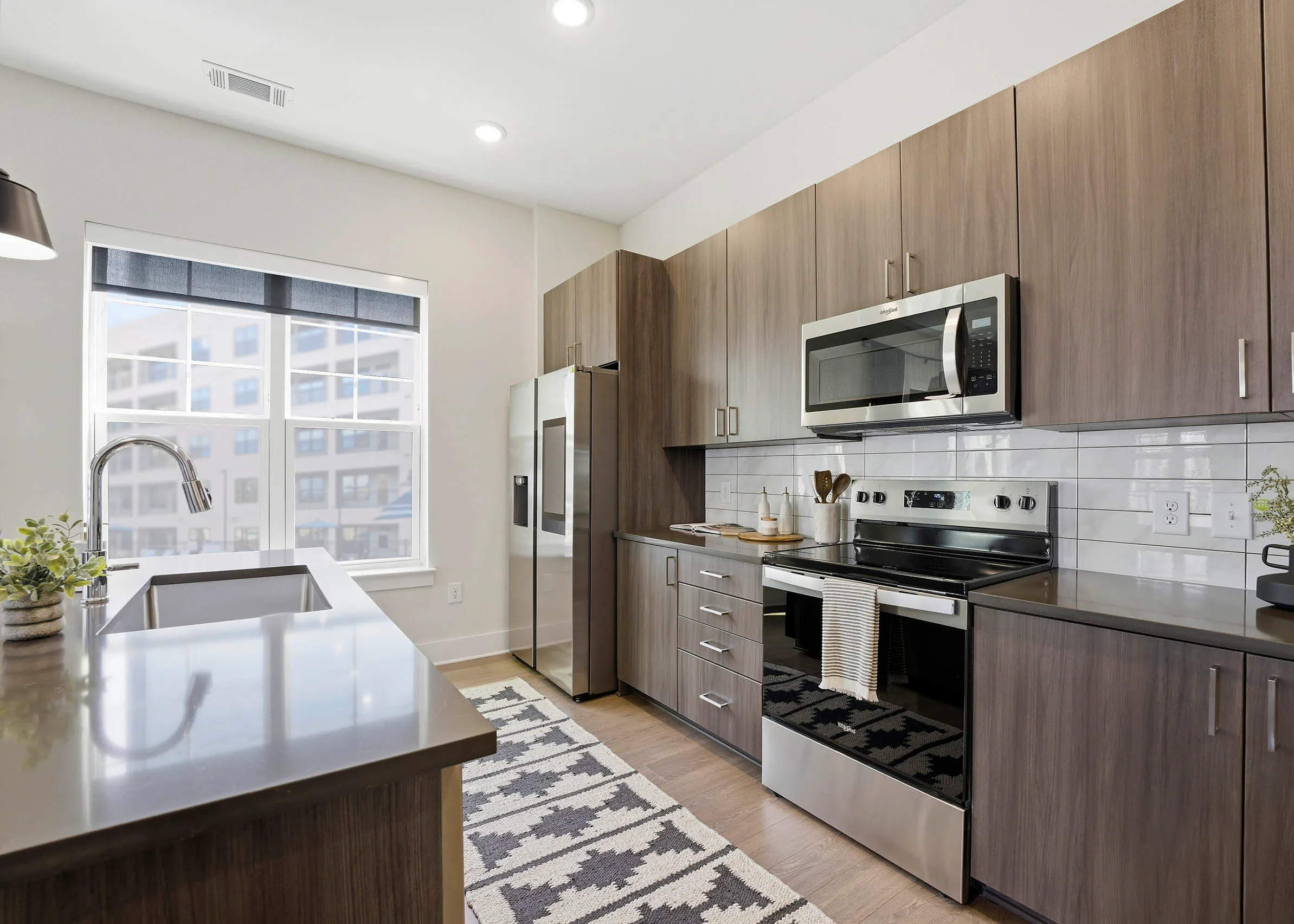 Kitchen with brown wood cabinetry, stainless steel appliances, brown island with sink, patterned cream and black rug, and large two pane window