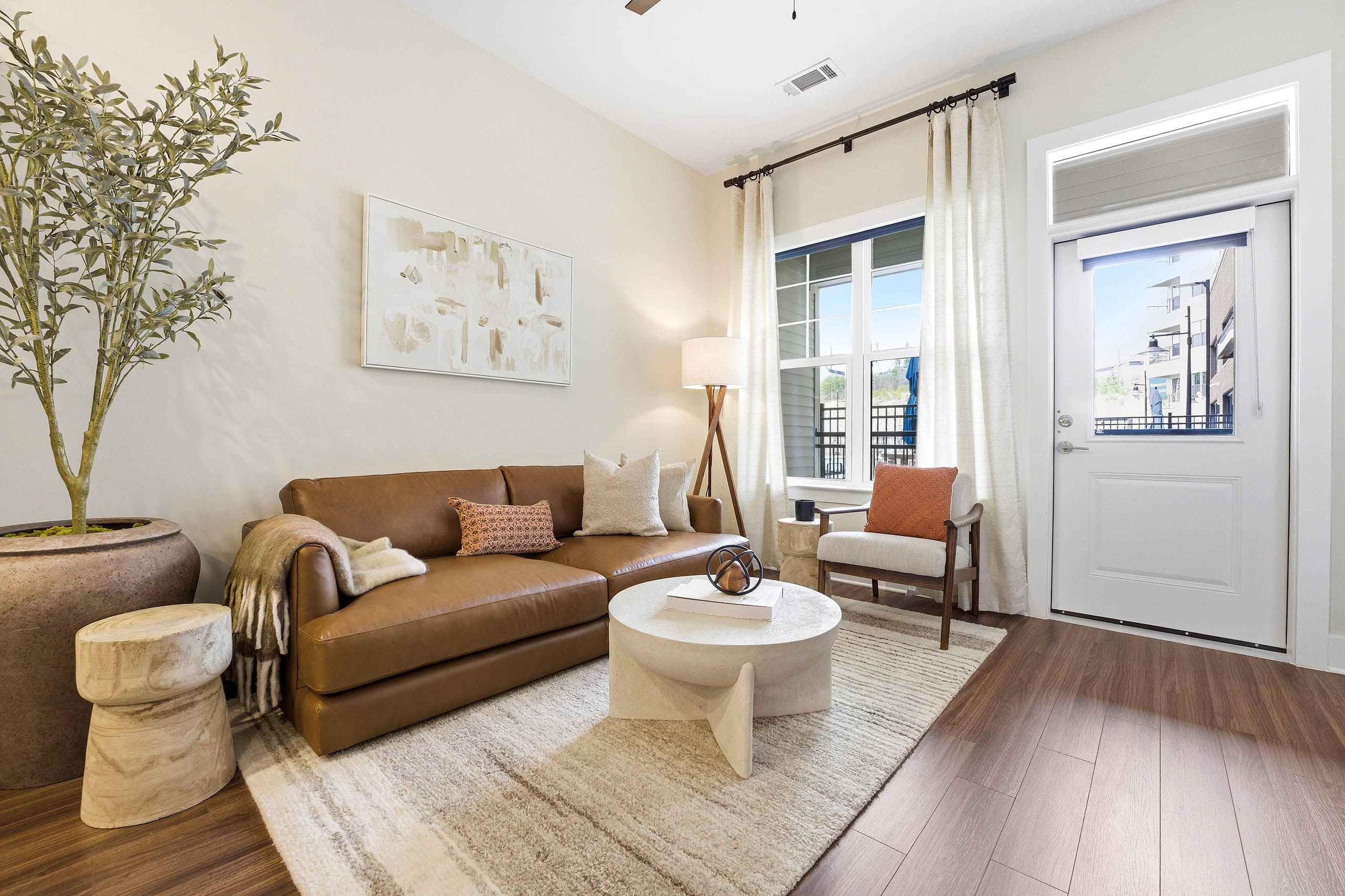 Bright living area with wood plank floors, brown leather couch, round cream coffee table, cream rug, corner lamp, large windows, and door to outside