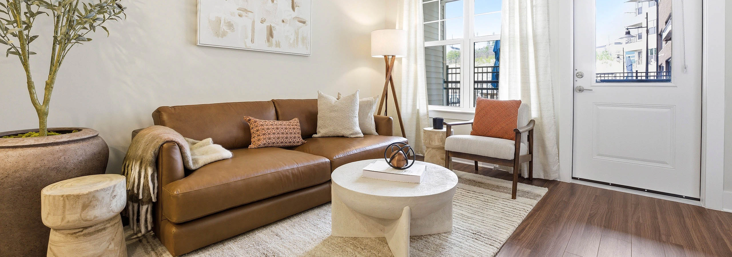 Bright living area with wood plank floors, brown leather couch, round cream coffee table, cream rug, corner lamp, large windows, and door to outside
