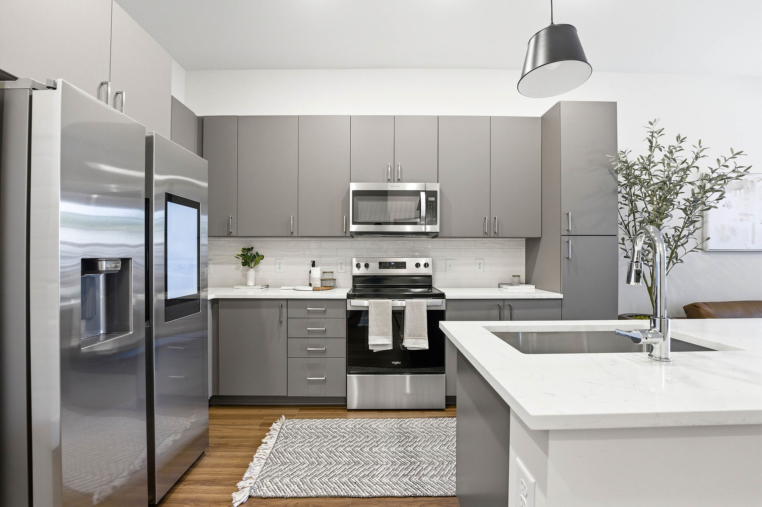Modern kitchen with gray cabinets, stainless steel appliances, touchscreen refrigerator, white countertops and white island with a sink