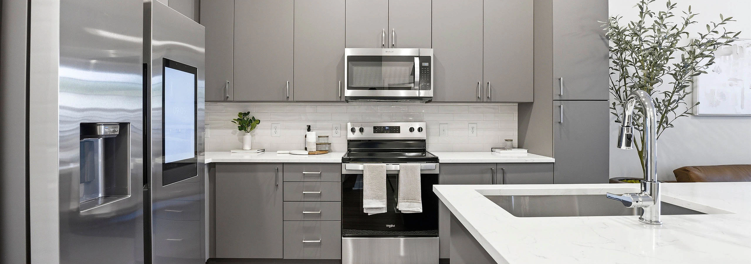 Modern kitchen with gray cabinets, stainless steel appliances, touchscreen refrigerator, white countertops and white island with a sink