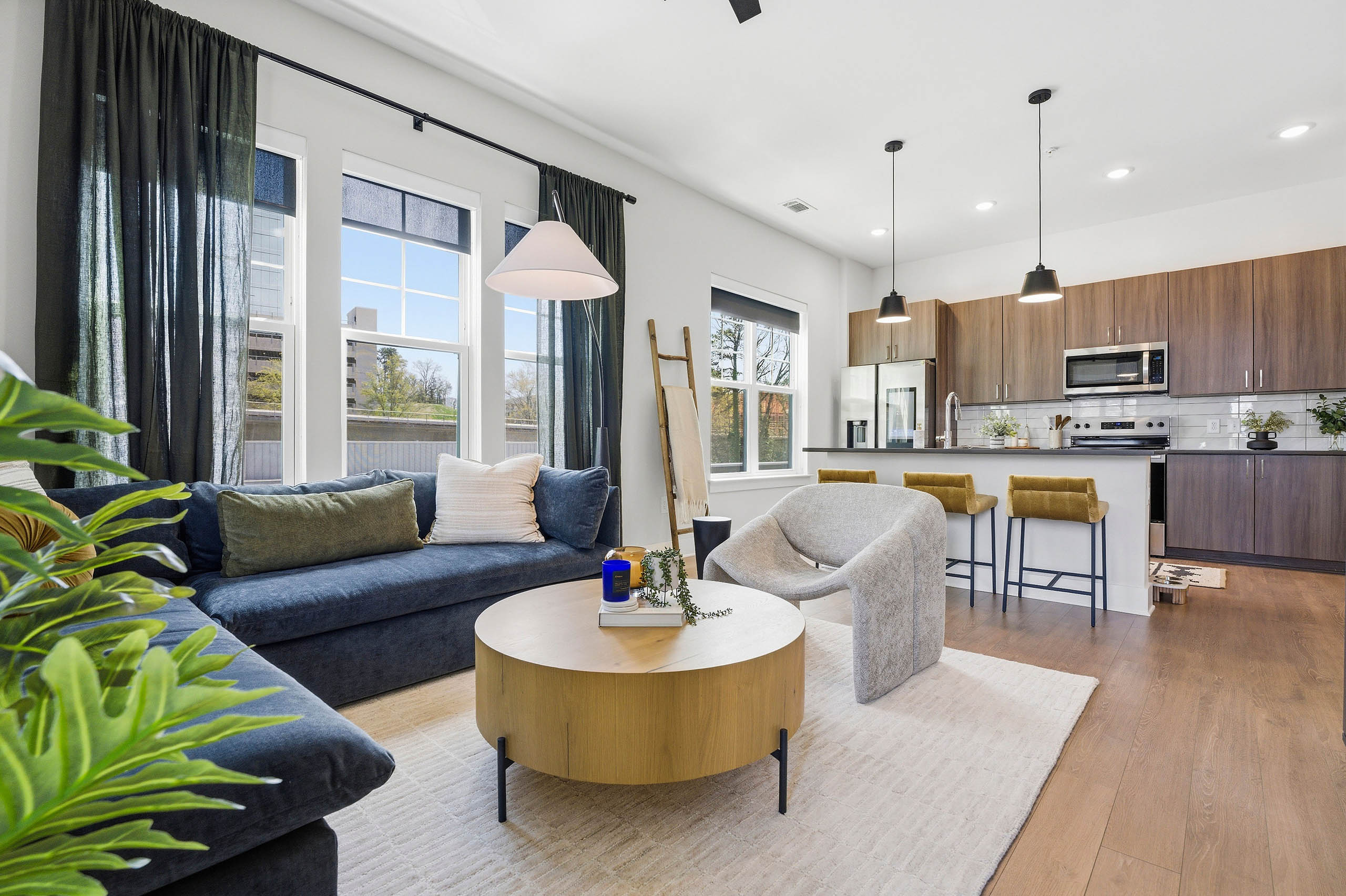 Open plan living room and kitchen with blue sectional, round wood coffee table, light rug, yellow bar stools, and large windows with dark curtains