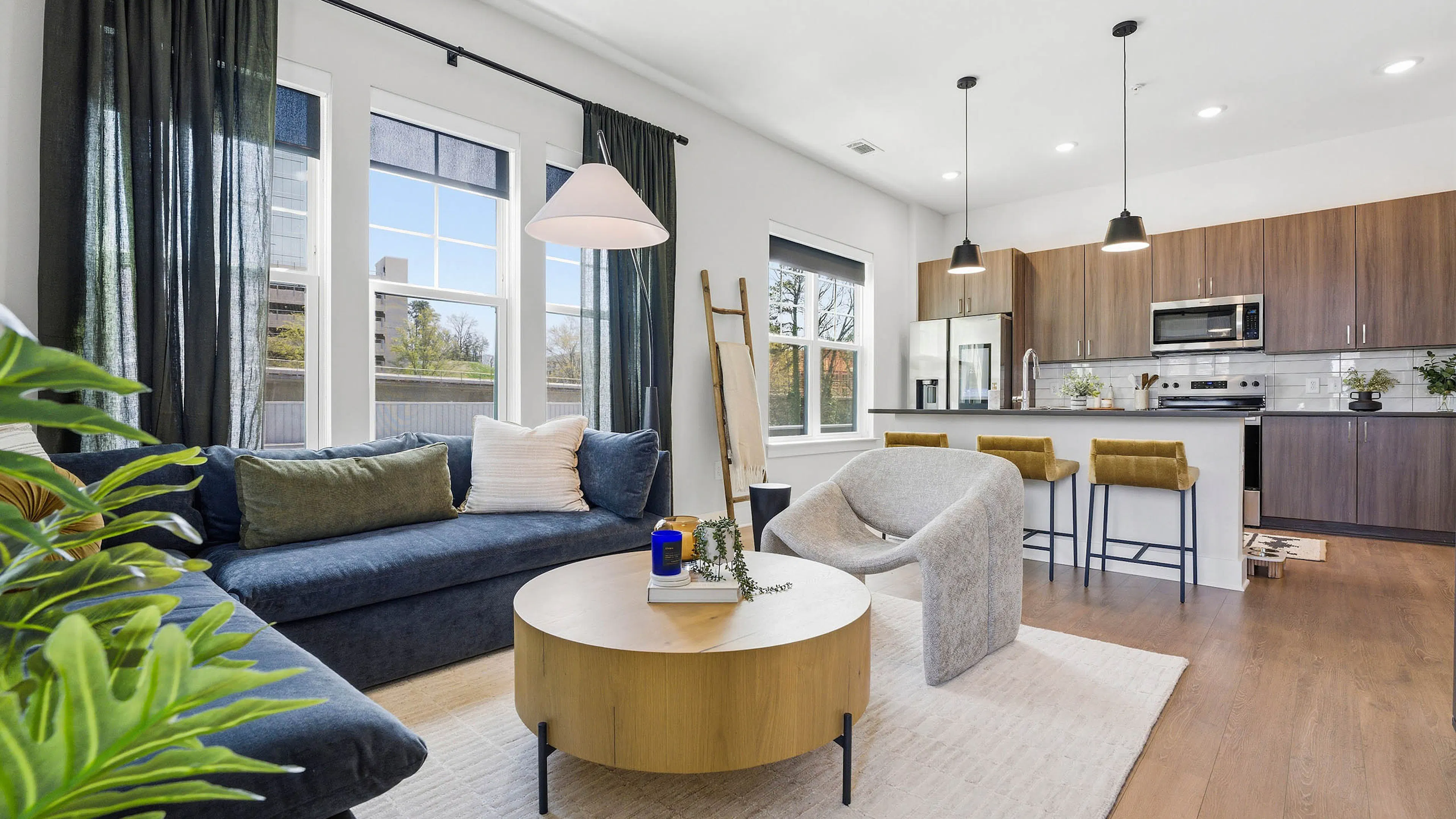 Open plan living room and kitchen with blue sectional, round wood coffee table, light rug, yellow bar stools, and large windows with dark curtains