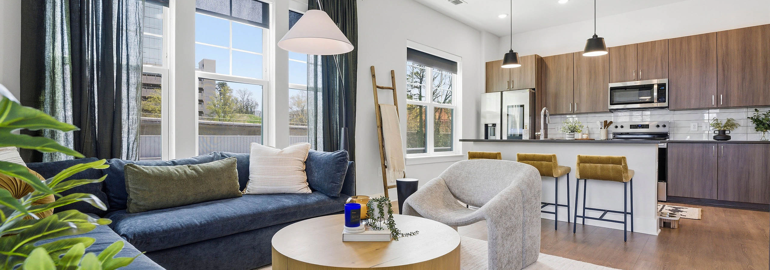 Open plan living room and kitchen with blue sectional, round wood coffee table, light rug, yellow bar stools, and large windows with dark curtains