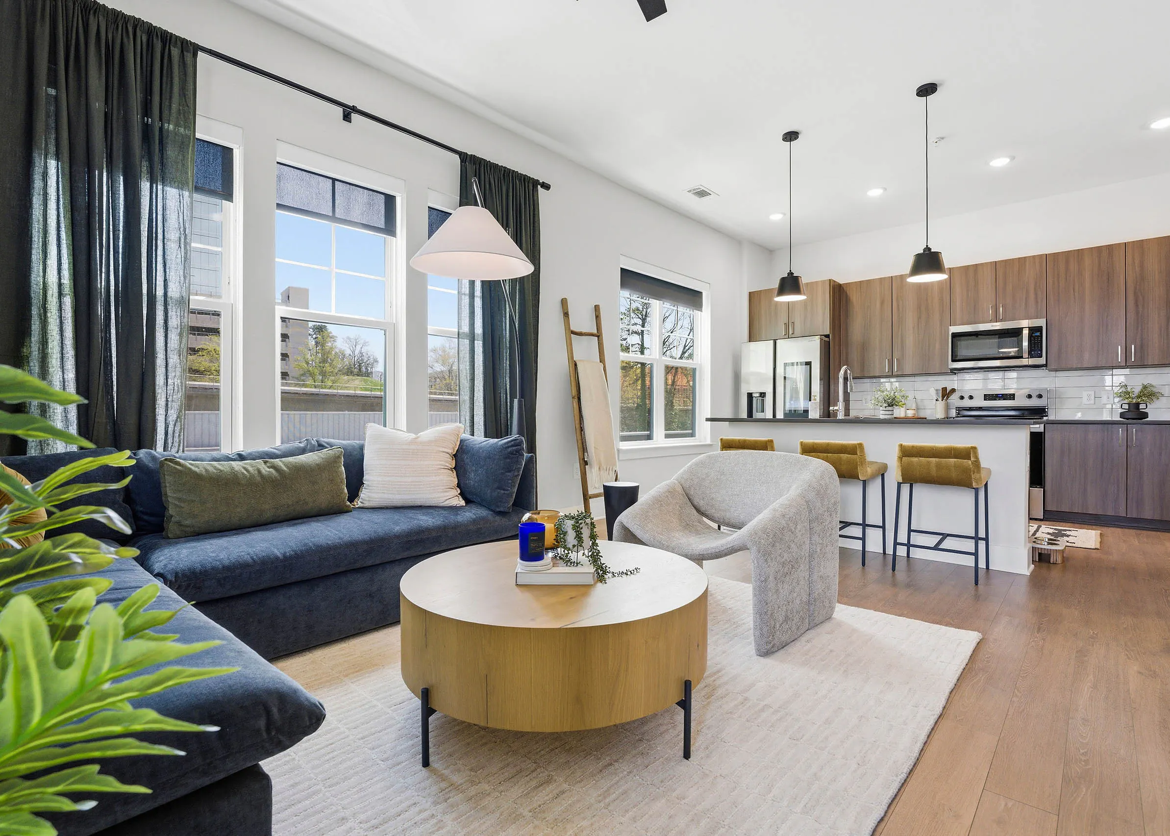 Open plan living room and kitchen with blue sectional, round wood coffee table, light rug, yellow bar stools, and large windows with dark curtains