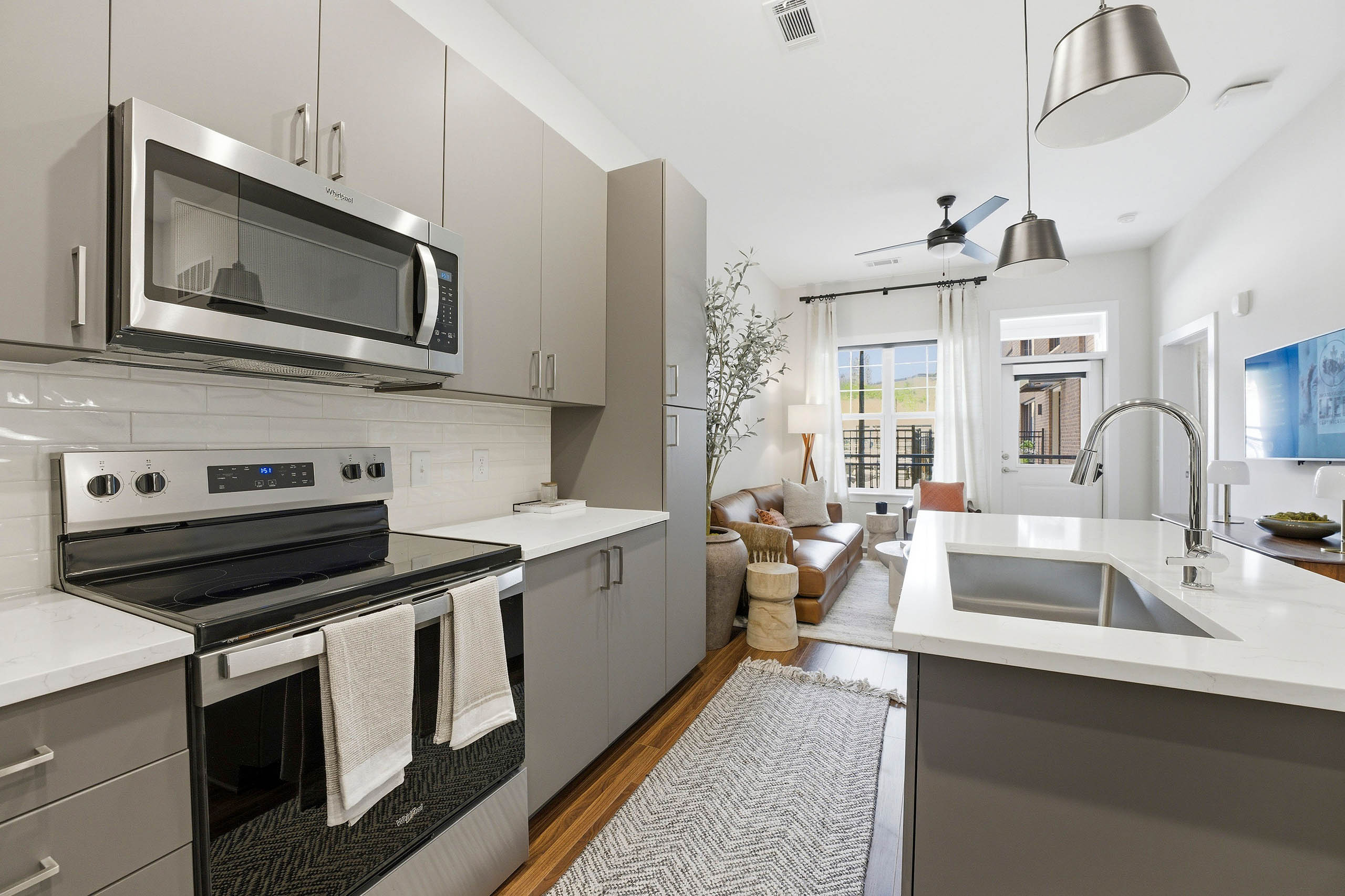 Modern kitchen with gray cabinets, stainless steel appliances, white island, opening to bright living area with brown couch, cream rug, plant, and coffee table