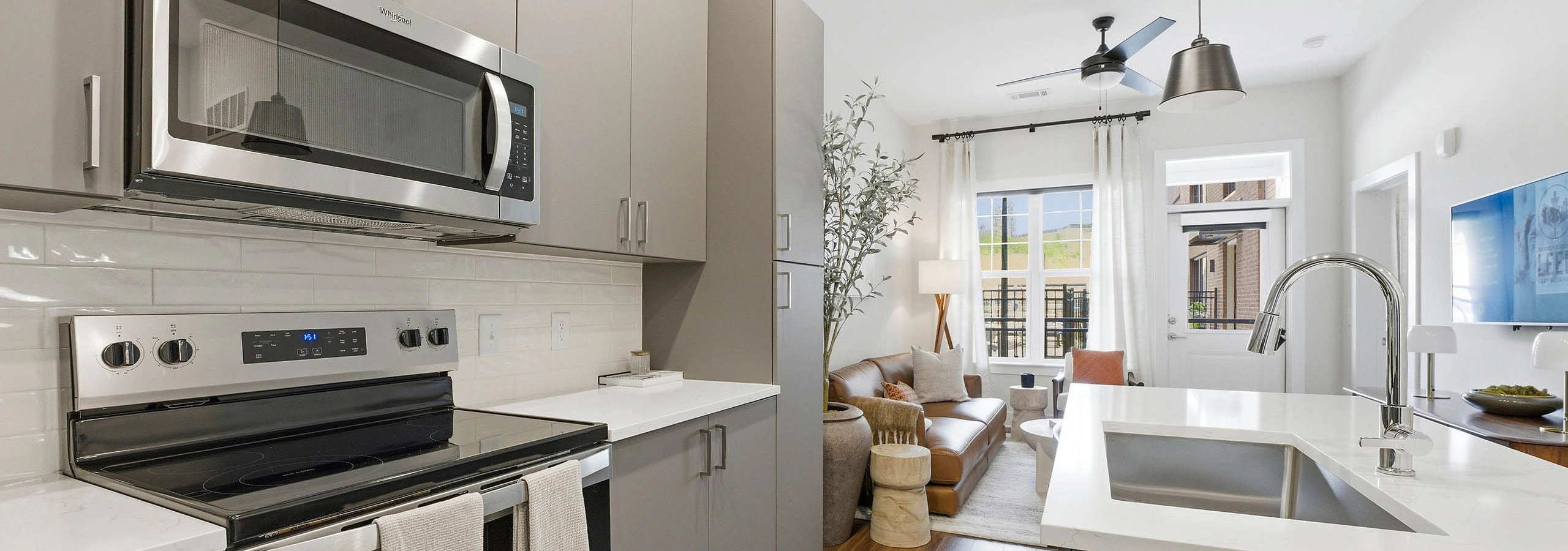 Modern kitchen with gray cabinets, stainless steel appliances, white island, opening to bright living area with brown couch, cream rug, plant, and coffee table