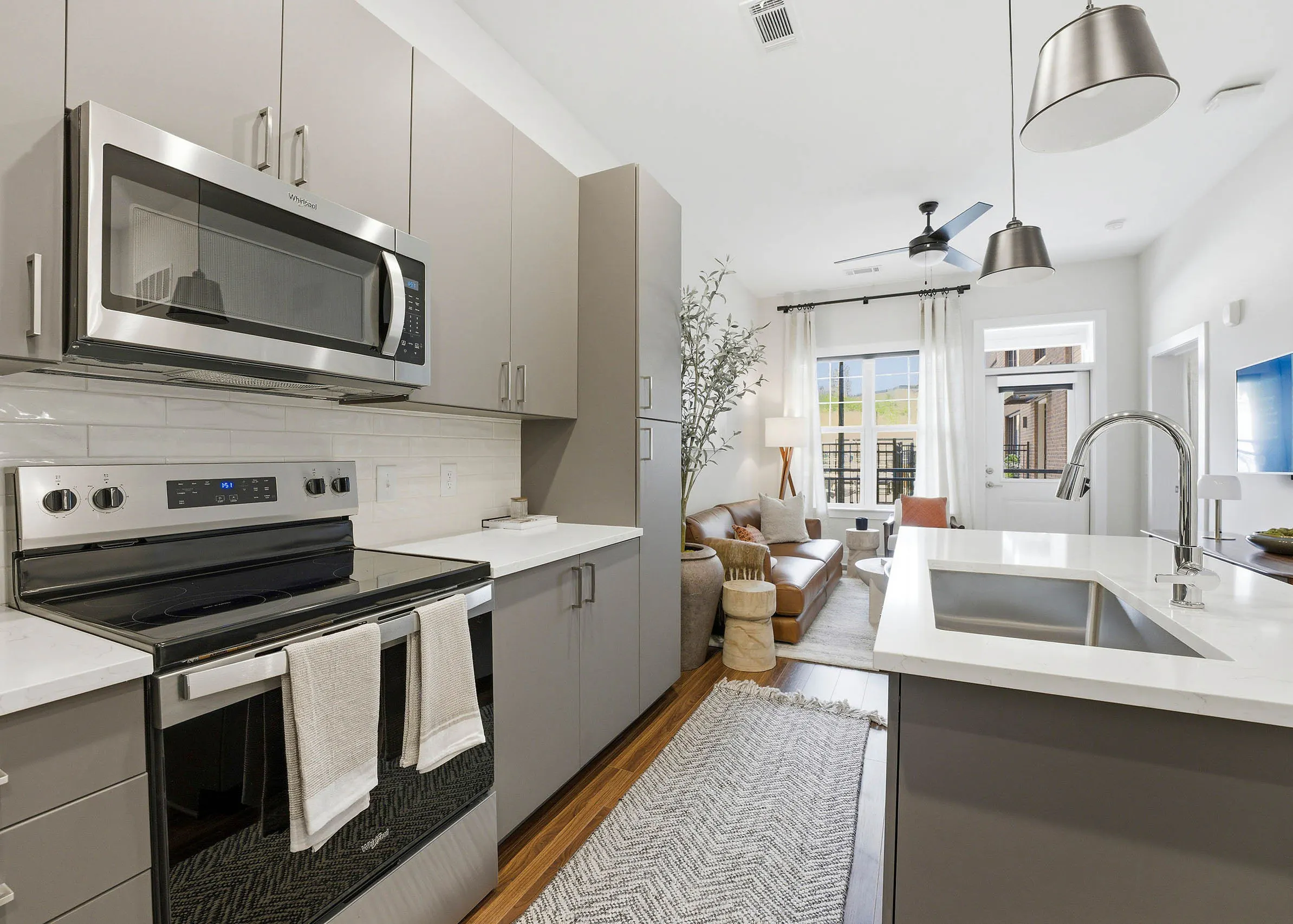 Modern kitchen with gray cabinets, stainless steel appliances, white island, opening to bright living area with brown couch, cream rug, plant, and coffee table
