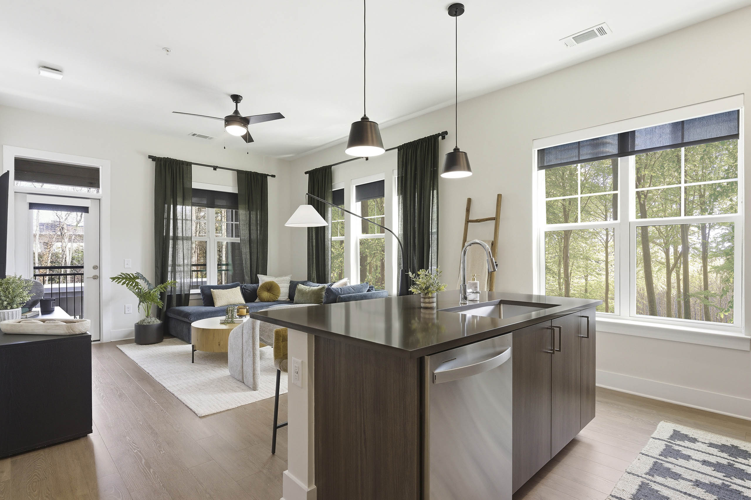 Bright kitchen and living area with brown island and sink, stainless steel dishwasher, blue sectional, wood coffee table, light rug, large windows