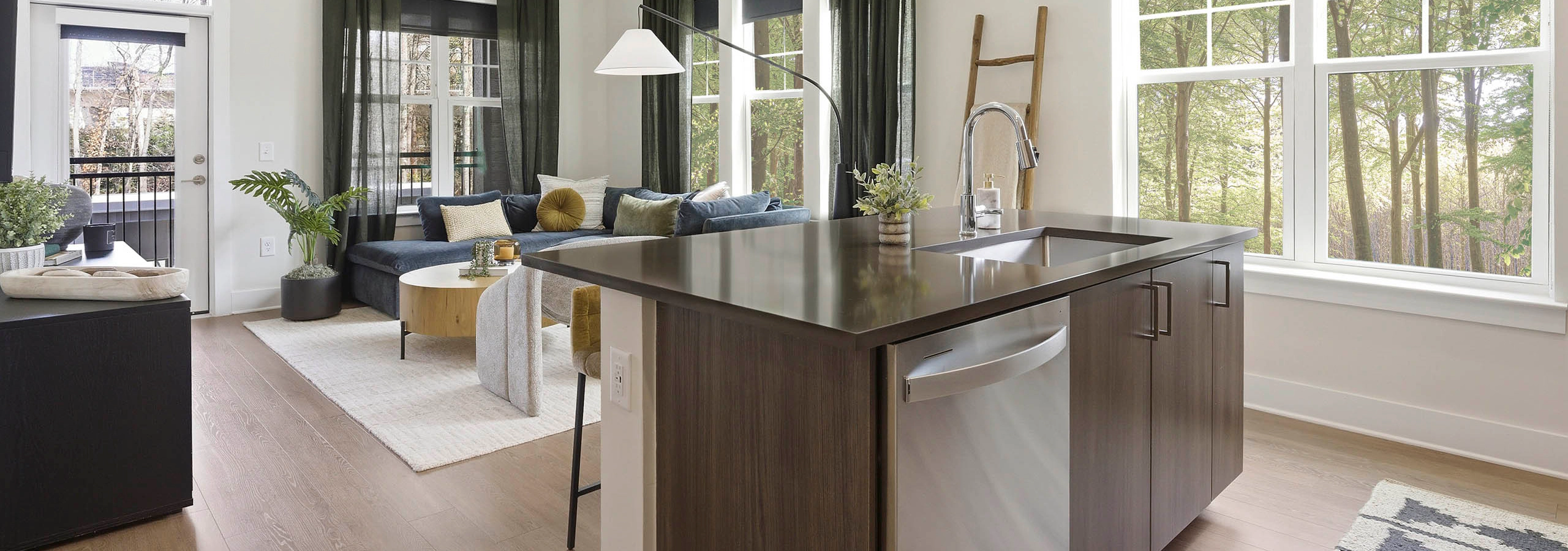 Bright kitchen and living area with brown island and sink, stainless steel dishwasher, blue sectional, wood coffee table, light rug, large windows