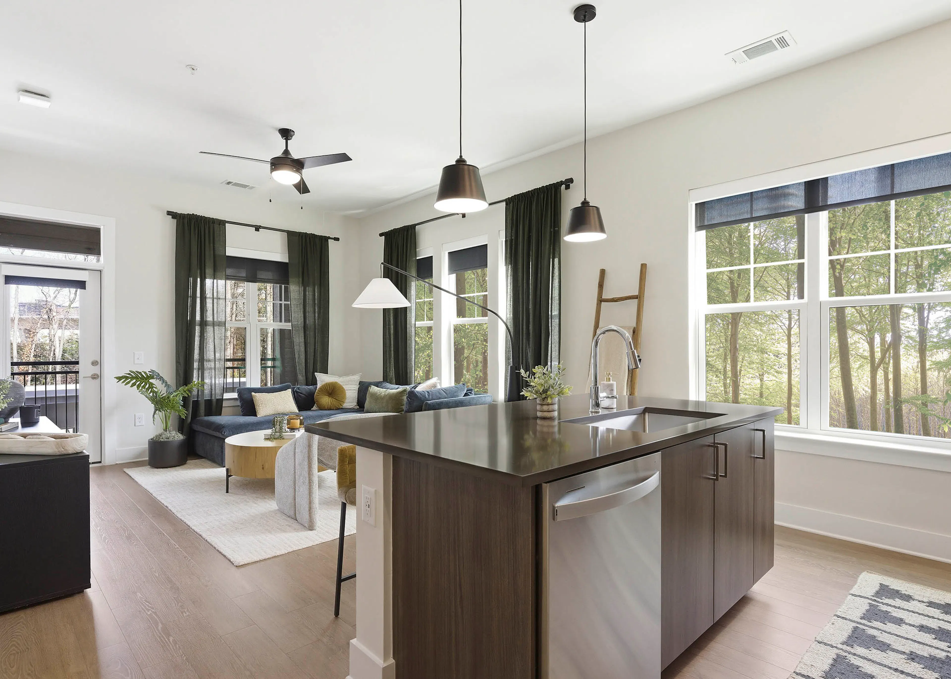 Bright kitchen and living area with brown island and sink, stainless steel dishwasher, blue sectional, wood coffee table, light rug, large windows
