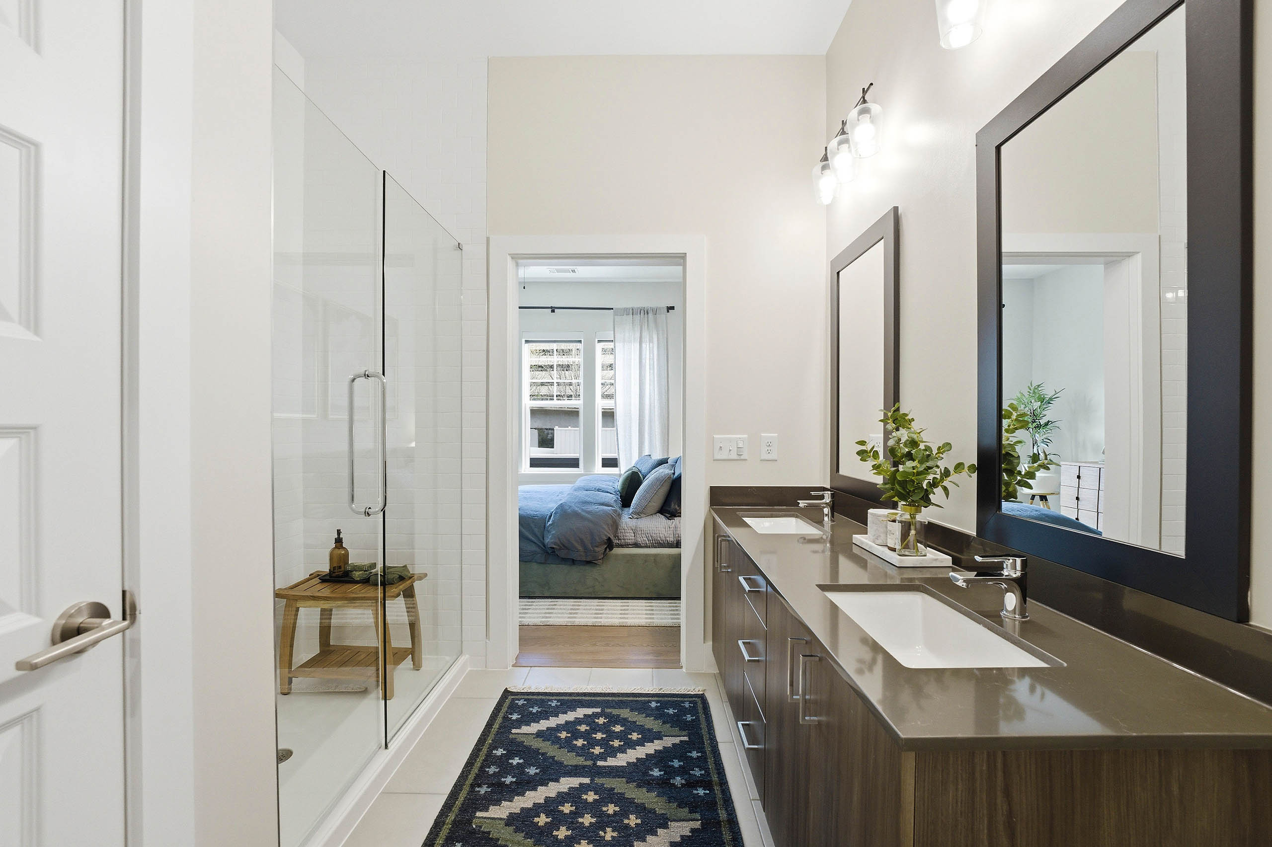 Bathroom with glass shower doors, brown double vanity unit with mirrors, white tiled floors, patterned rug, and a bedroom visible through the doorway