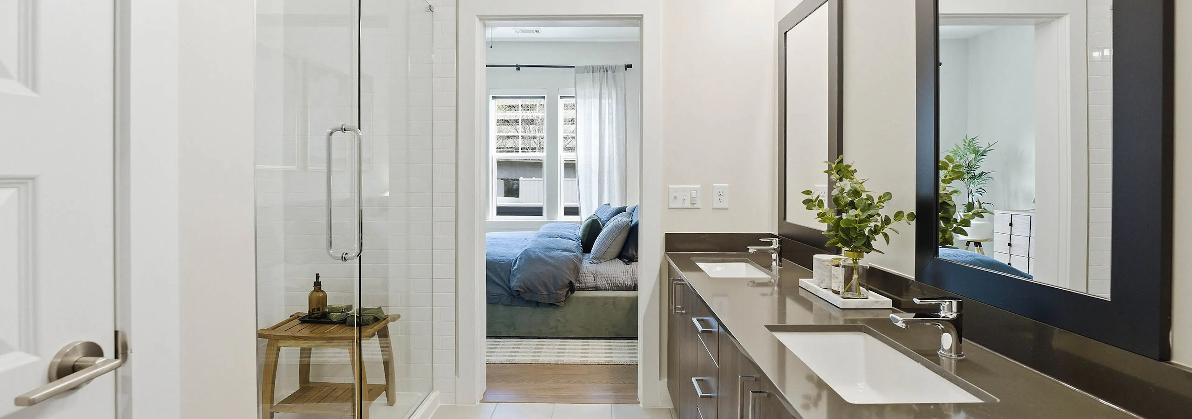 Bathroom with glass shower doors, brown double vanity unit with mirrors, white tiled floors, patterned rug, and a bedroom visible through the doorway