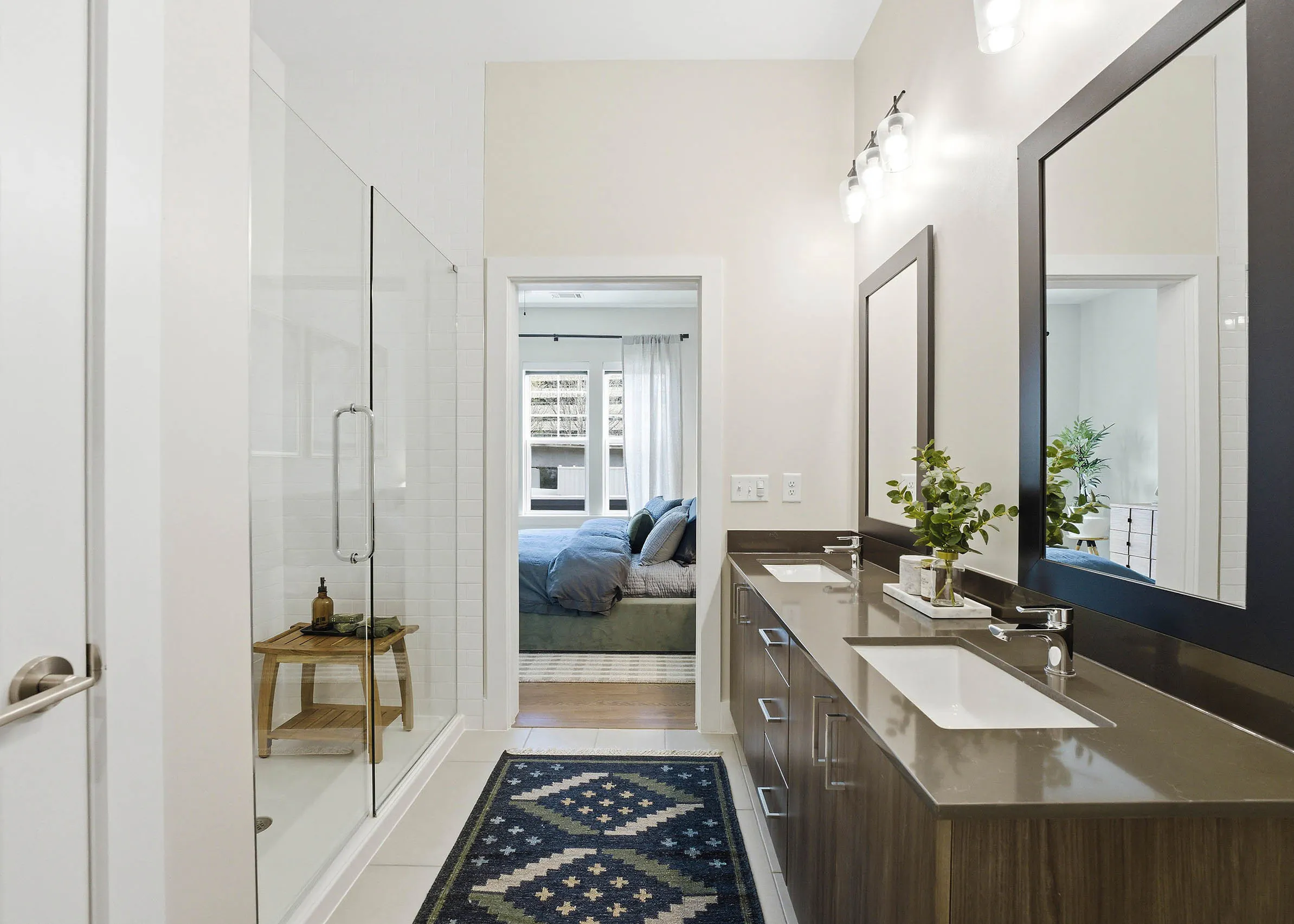 Bathroom with glass shower doors, brown double vanity unit with mirrors, white tiled floors, patterned rug, and a bedroom visible through the doorway