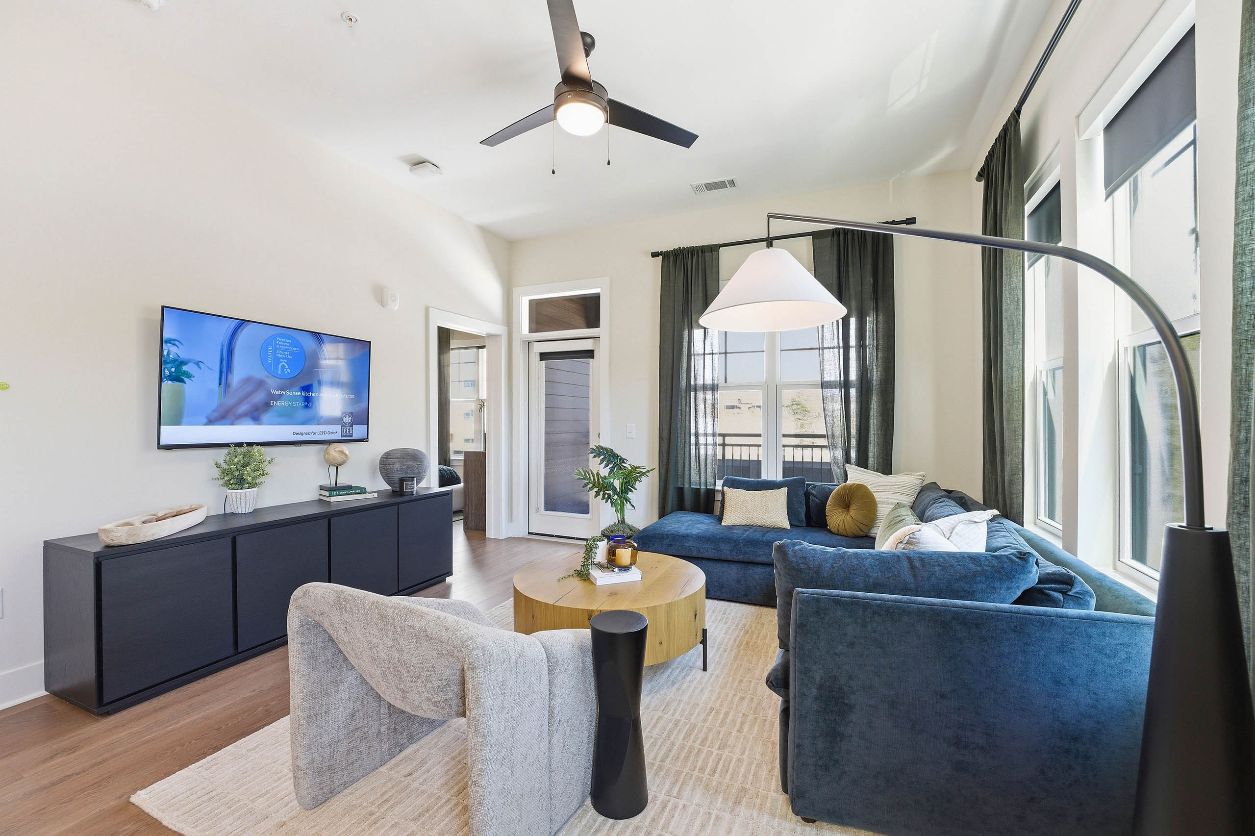 Open plan living room and kitchen with blue sectional, round wood coffee table, light rug, black console with wall-mounted TV and large windows