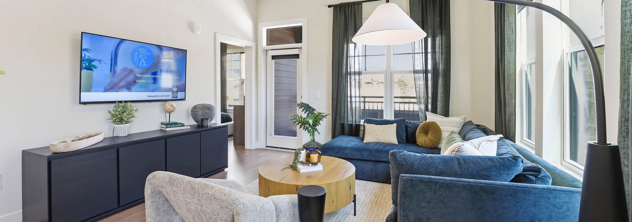 Open plan living room and kitchen with blue sectional, round wood coffee table, light rug, black console with wall-mounted TV and large windows