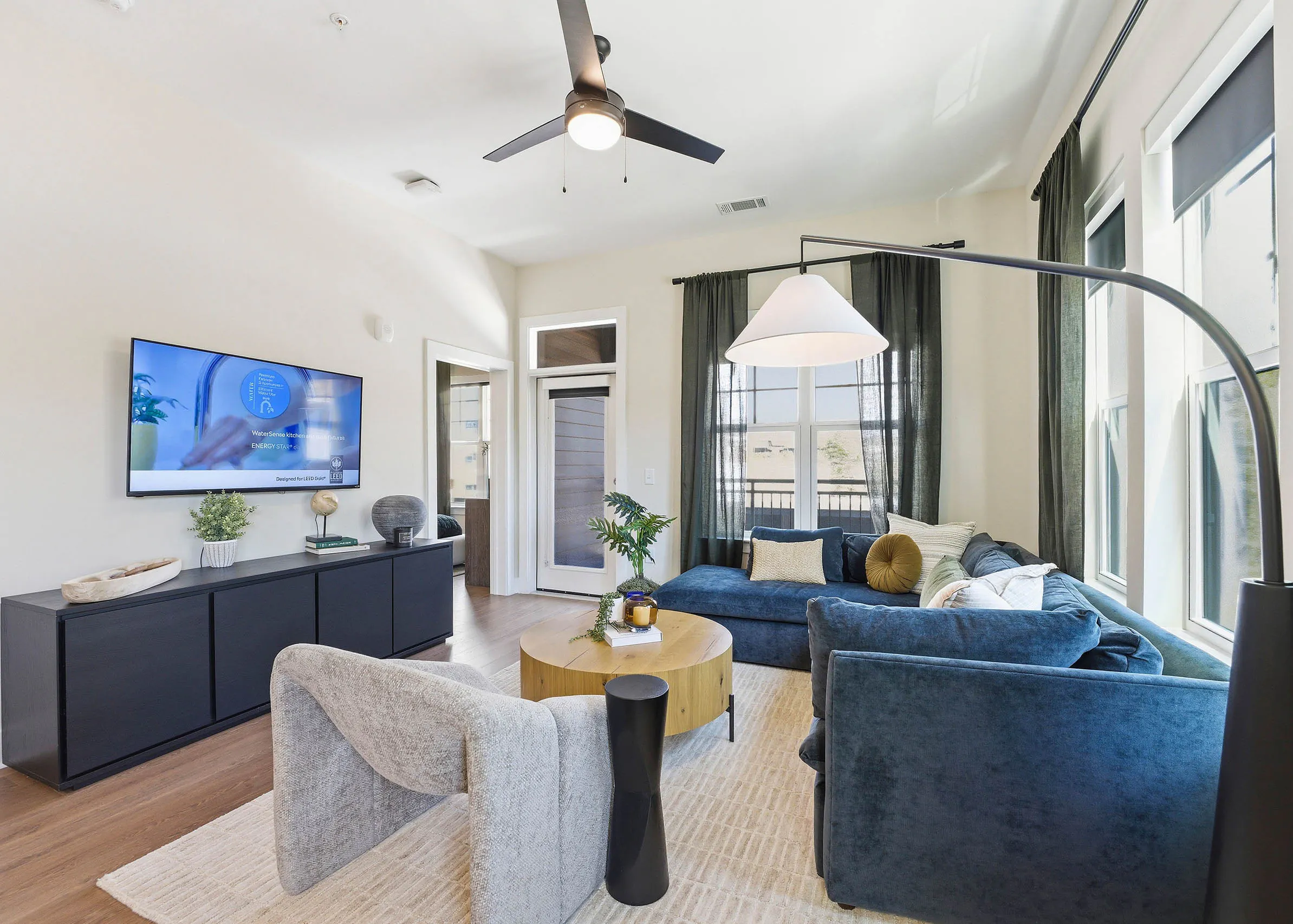 Open plan living room and kitchen with blue sectional, round wood coffee table, light rug, black console with wall-mounted TV and large windows