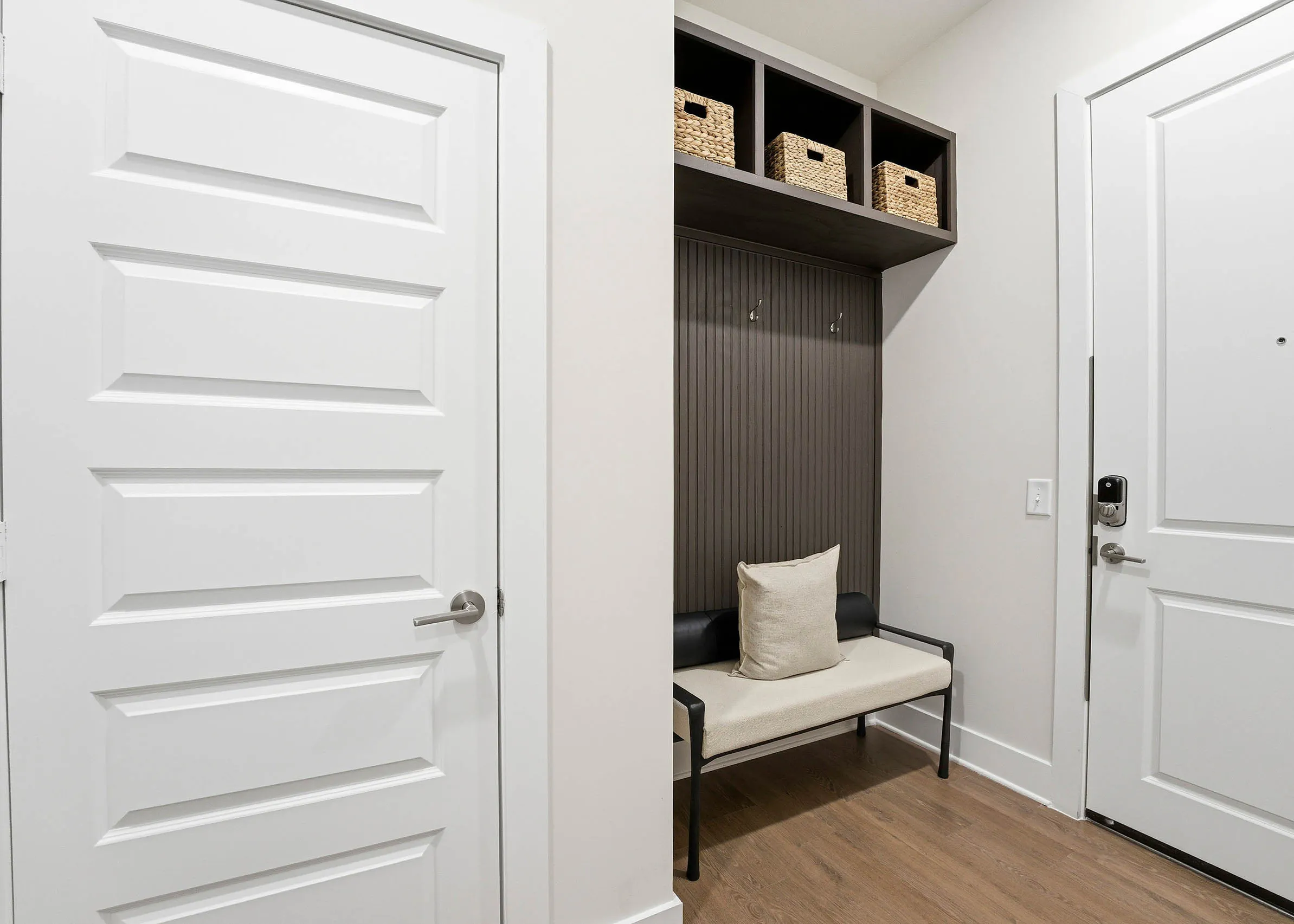 Back of apartment door; black seat with white pillow, storage with three straw boxes against brown wall, and a closed white door