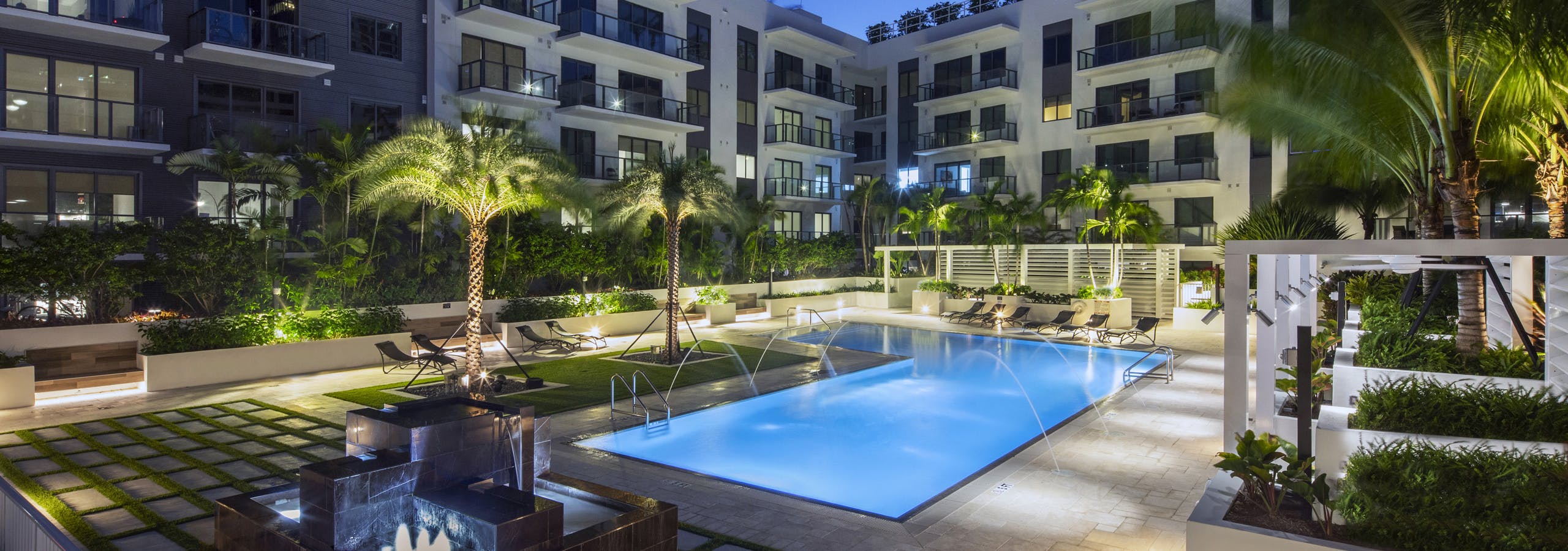 AMLI Midtown Miami courtyard pool lit up at night with arching water streams surrounded by cabanas and lush landscape