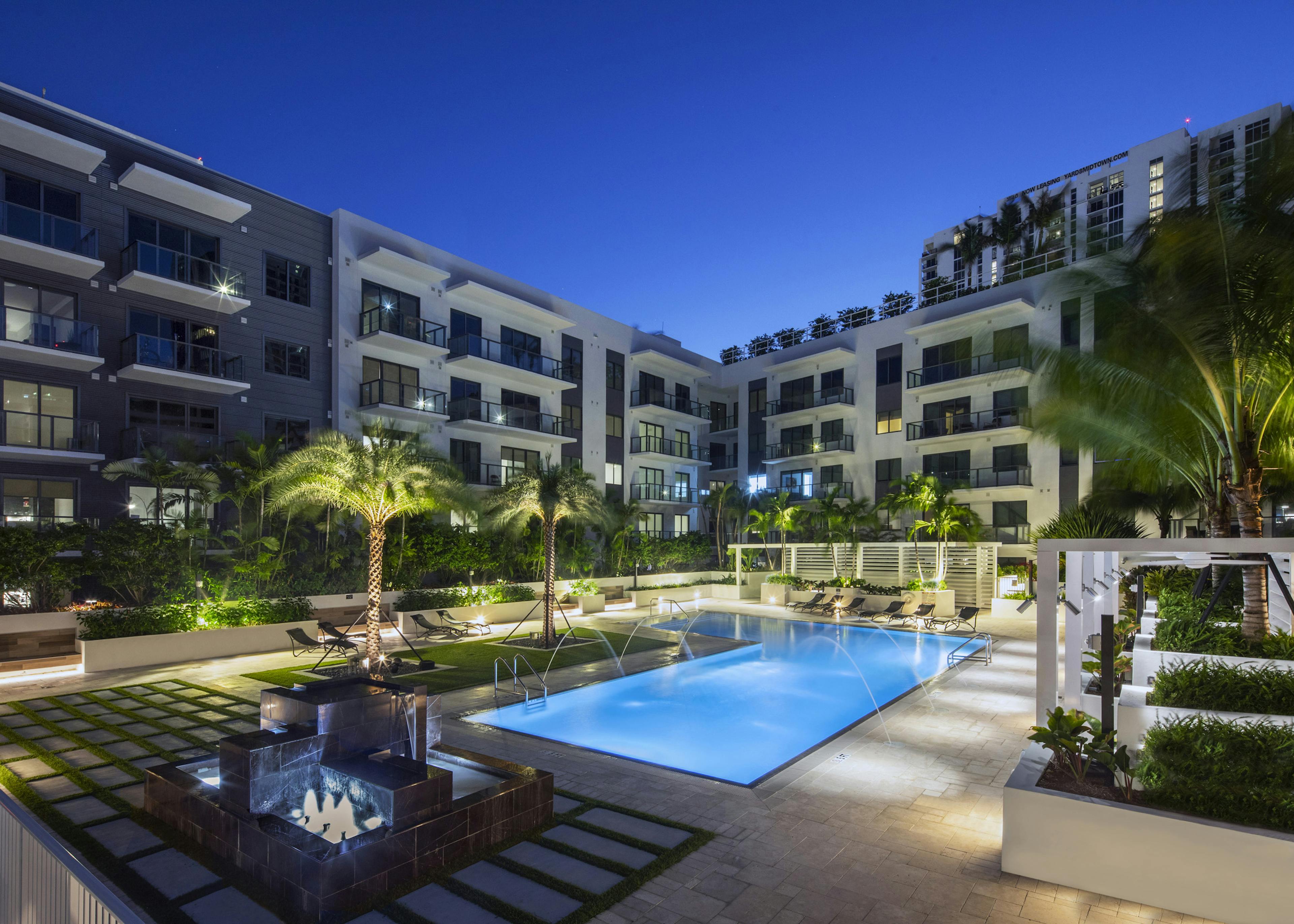 AMLI Midtown Miami courtyard pool lit up at night with arching water streams surrounded by cabanas and lush landscape