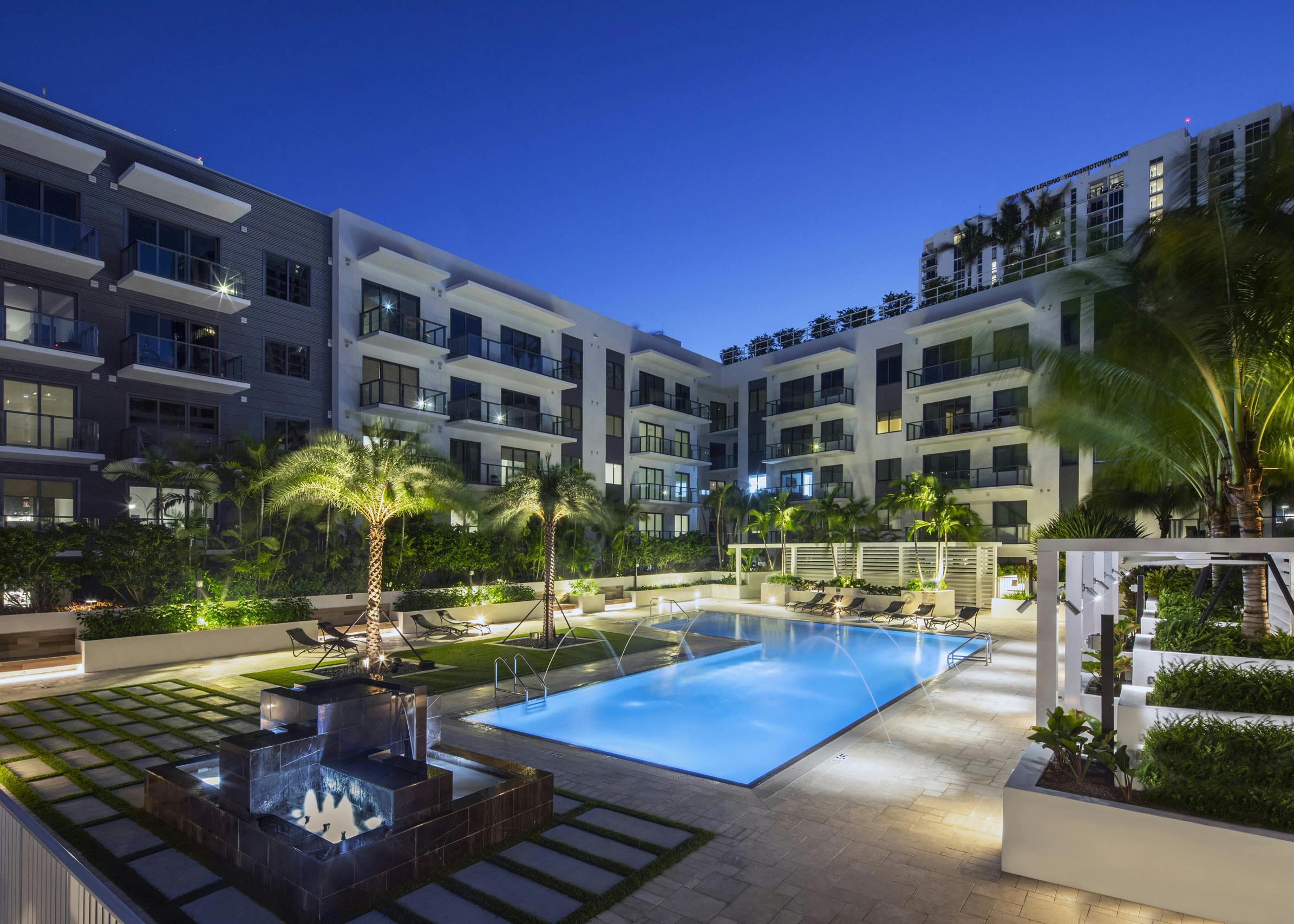 AMLI Midtown Miami courtyard pool lit up at night with arching water streams surrounded by cabanas and lush landscape