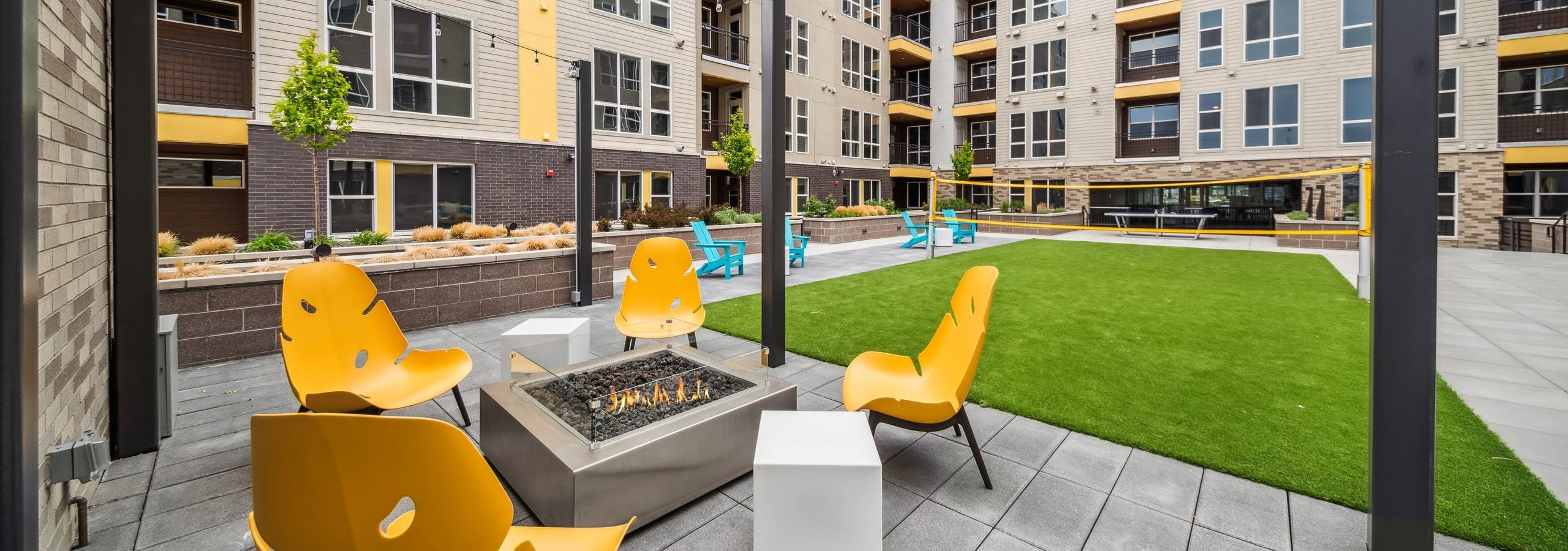 AMLI RiNo courtyard with a firepit surrounded by 4 yellow chairs with string lights above adjacent to a turf badminton court
