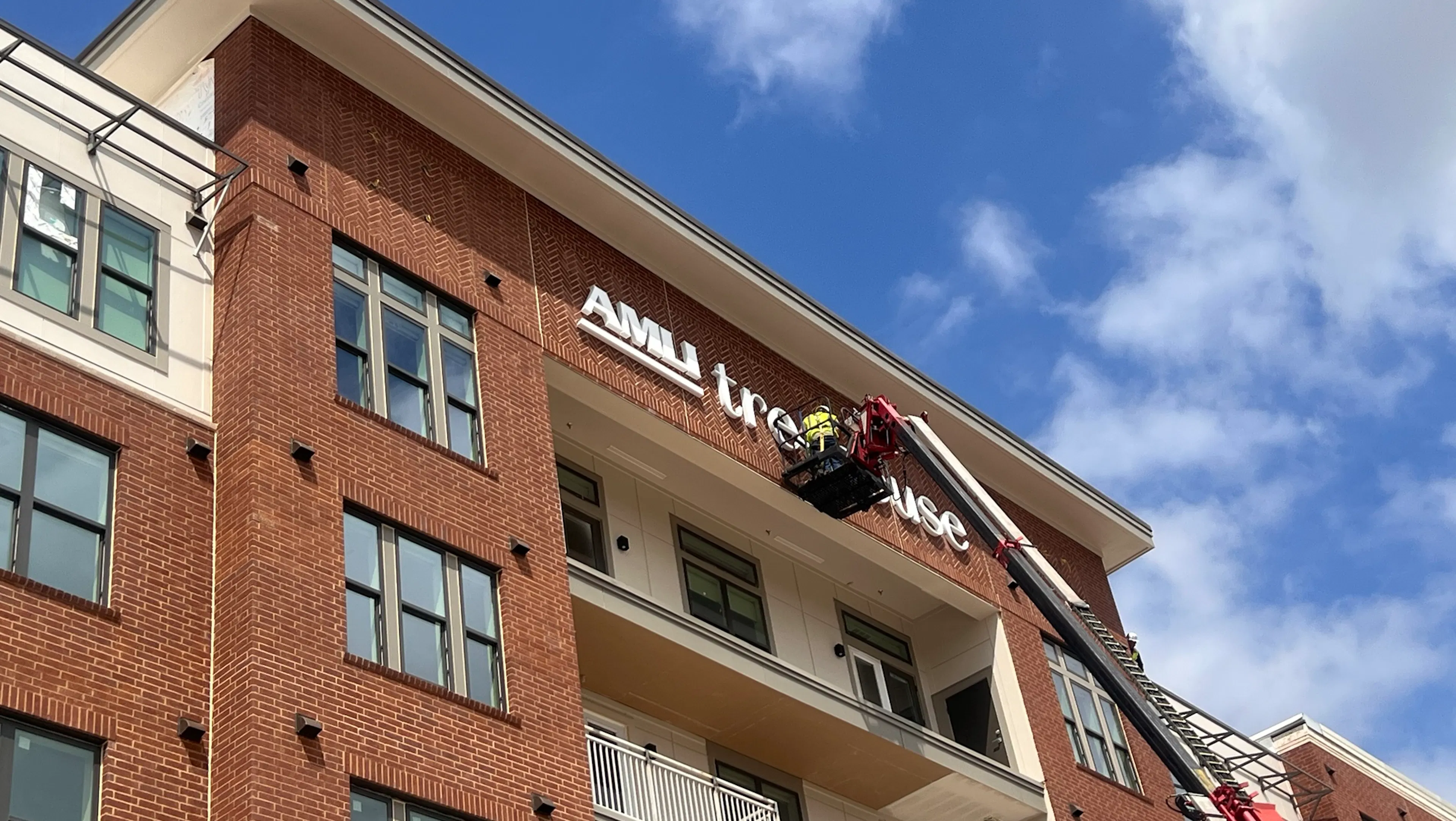 Exterior shot of an apartment building with “AMLI Treehouse” signage being installed by workers on a lift against a blue sky.