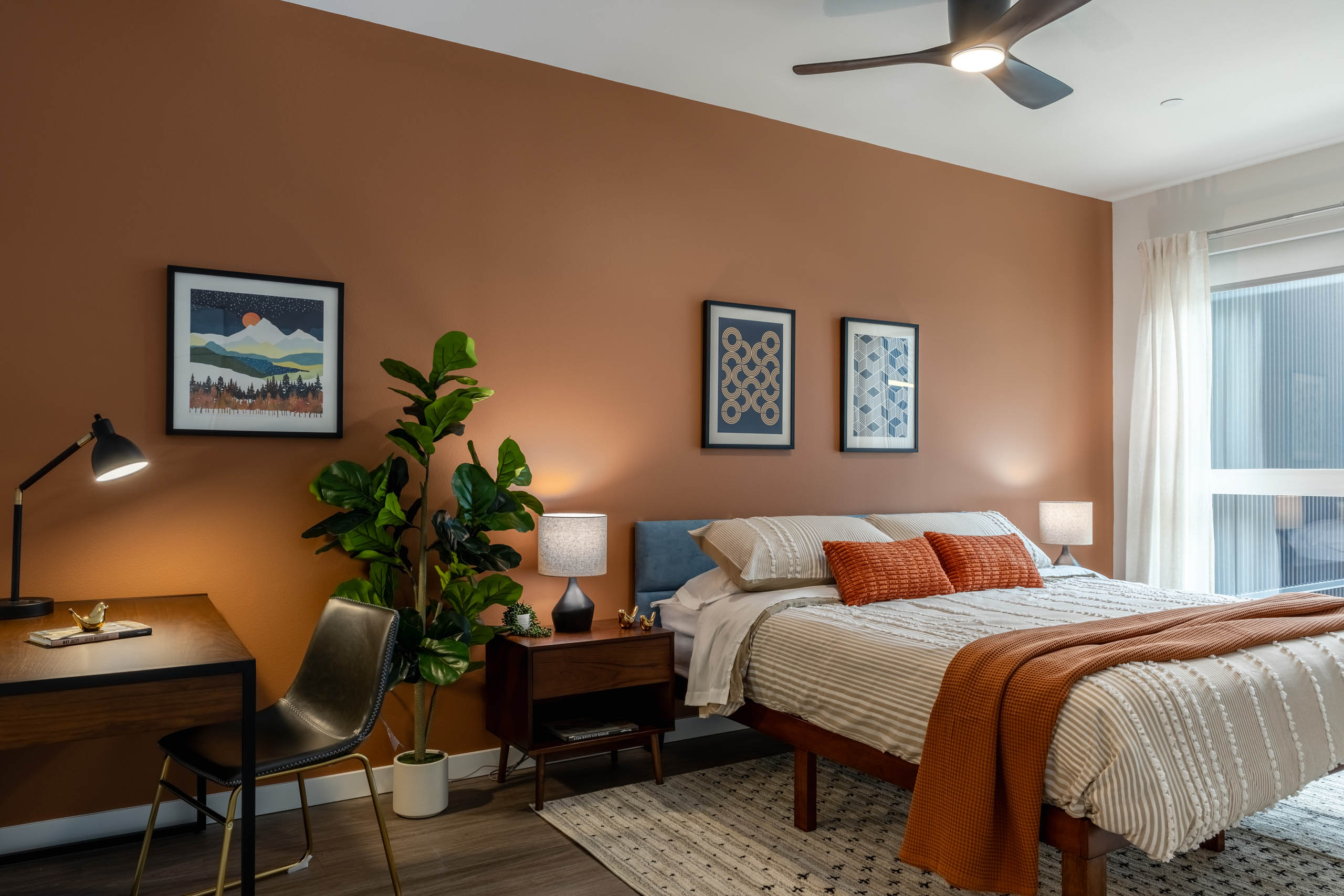 Large bedroom with orange walls, bed with wooden nightstand, and small wooden desk in the corner.
