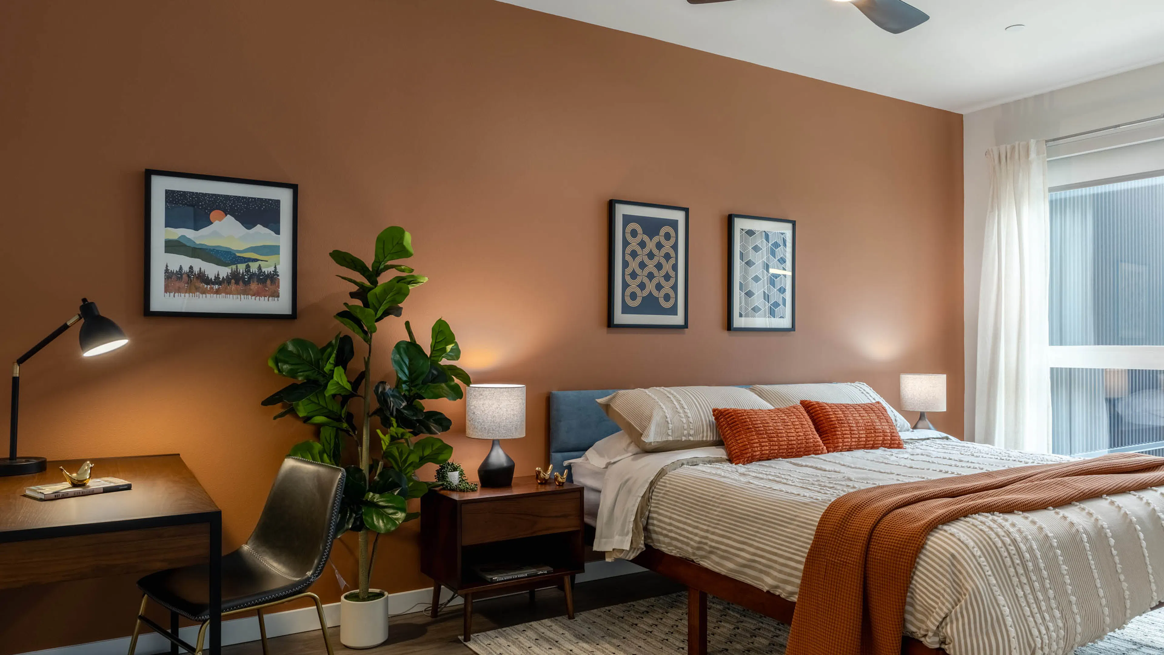 Large bedroom with orange walls, large bed with wooden nightstand and lamp. Small wooden desk in the corner next to a potted plant.