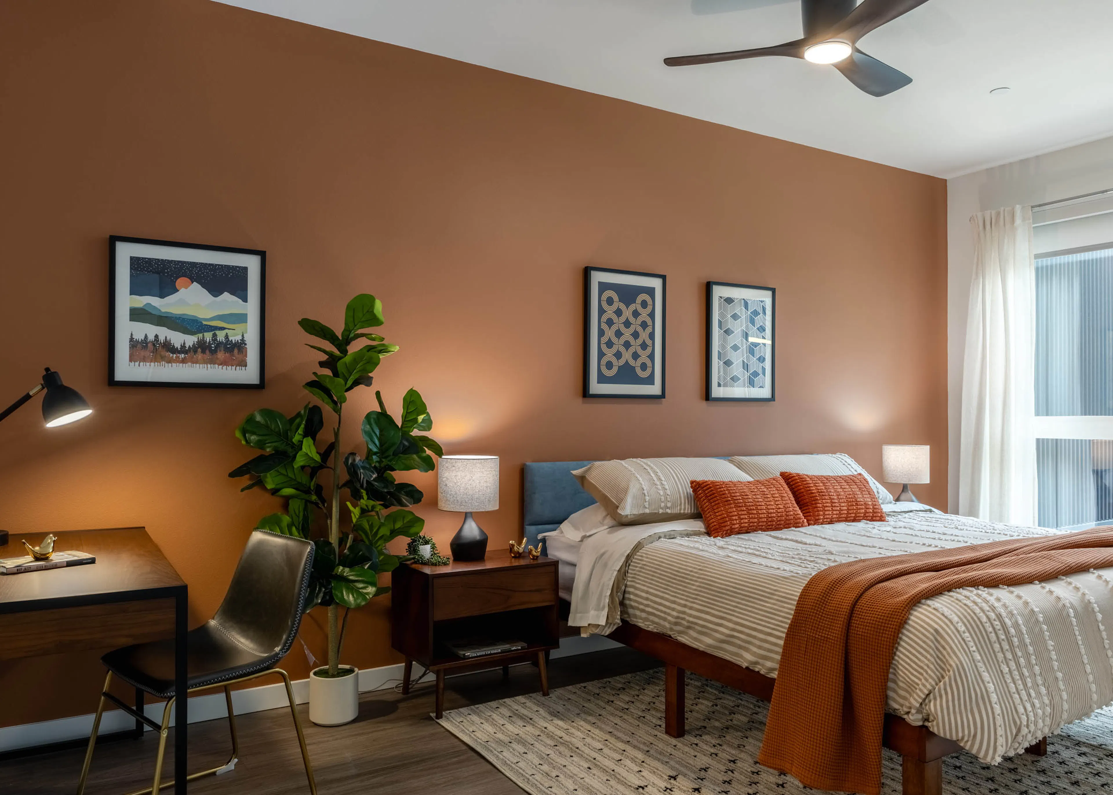 Large bedroom with orange walls, large bed with wooden nightstand and lamp. Small wooden desk in the corner next to a potted plant.