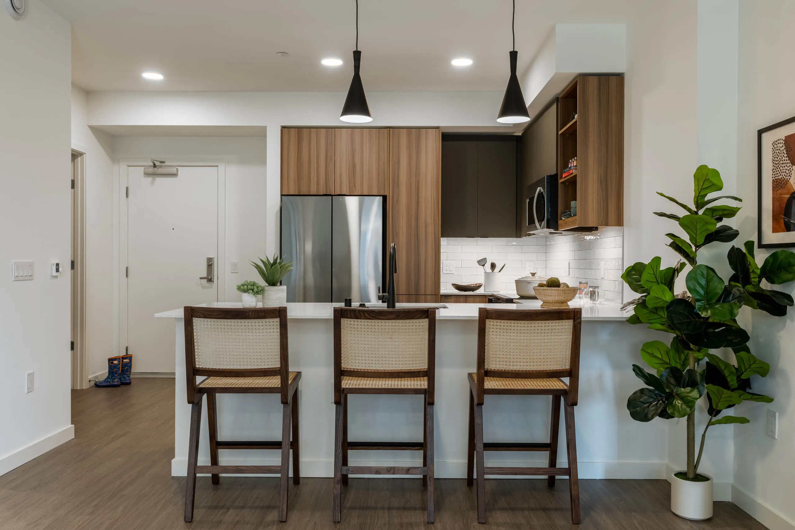 Kitchen with stainless steel appliances, wooden brown and grey cabinetry large white countertop and 3 highchairs and hanging spotlights above.