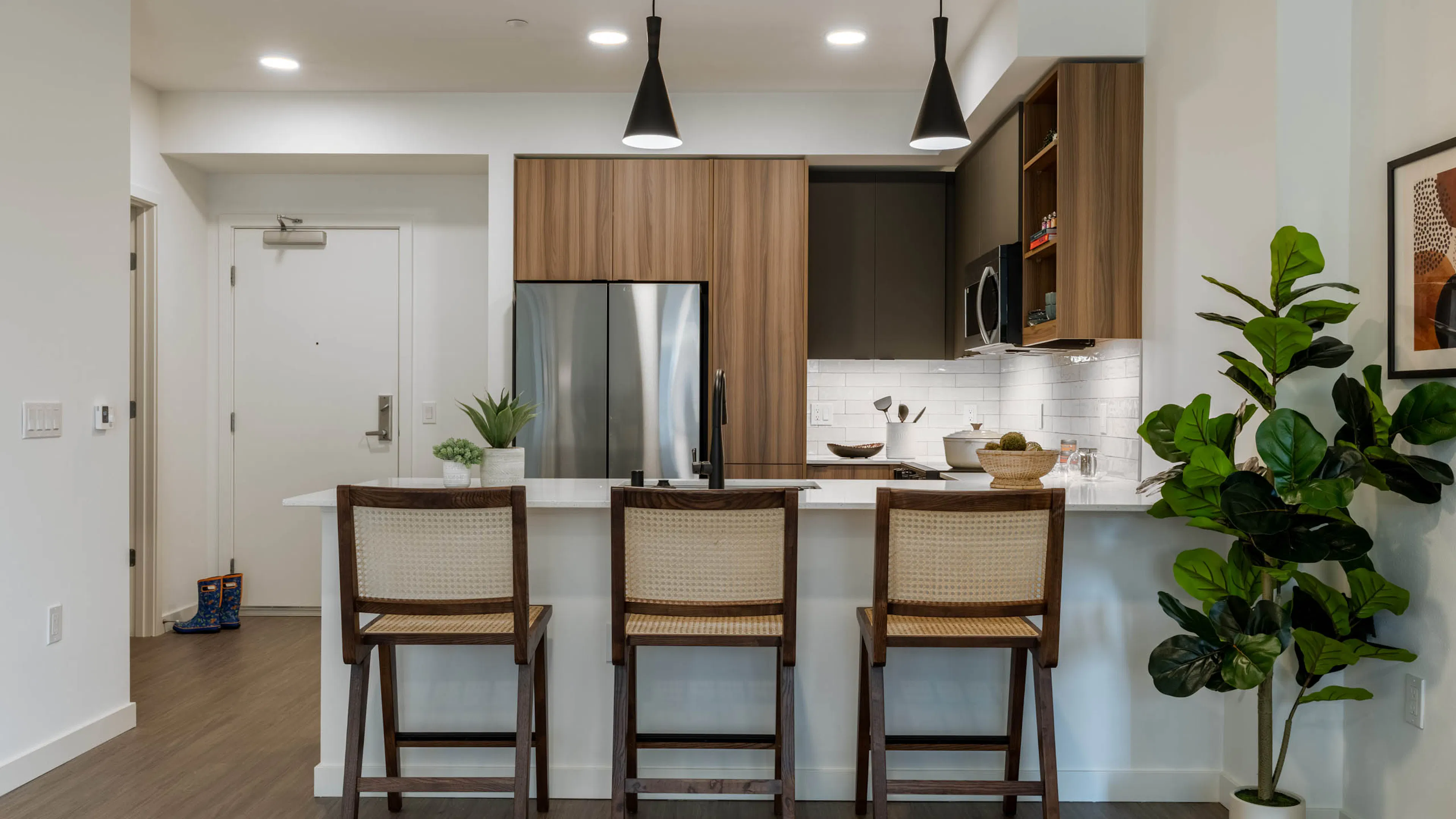 Kitchen with stainless steel appliances, wooden brown and grey cabinetry large white countertop and 3 highchairs and hanging spotlights above.