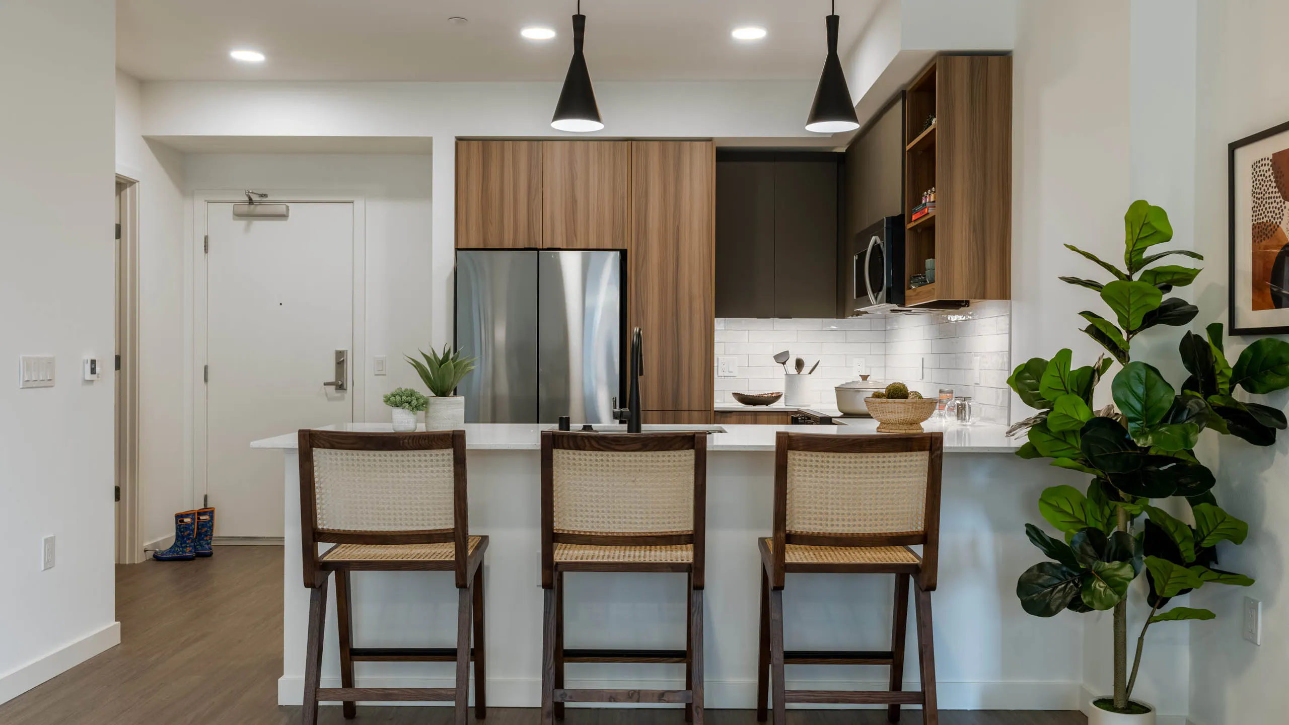 Kitchen with stainless steel appliances, wooden brown and grey cabinetry large white countertop and 3 highchairs and hanging spotlights above.