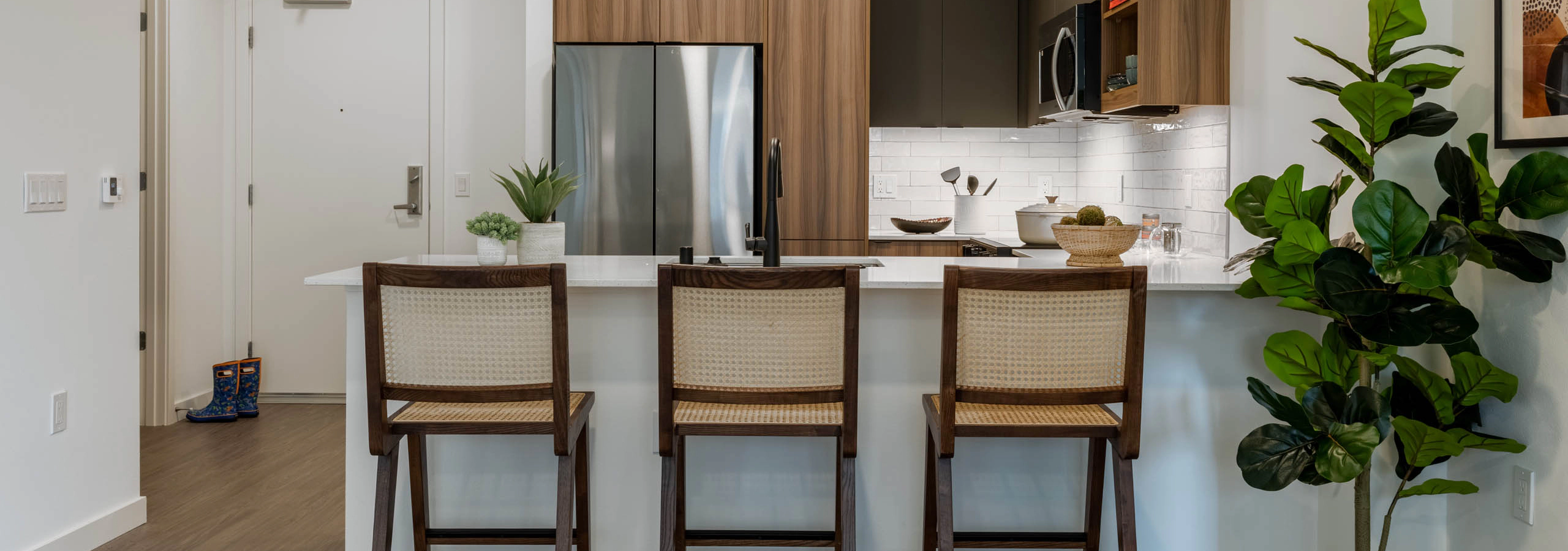Kitchen with stainless steel appliances, wooden brown and grey cabinetry large white countertop and 3 highchairs and hanging spotlights above.