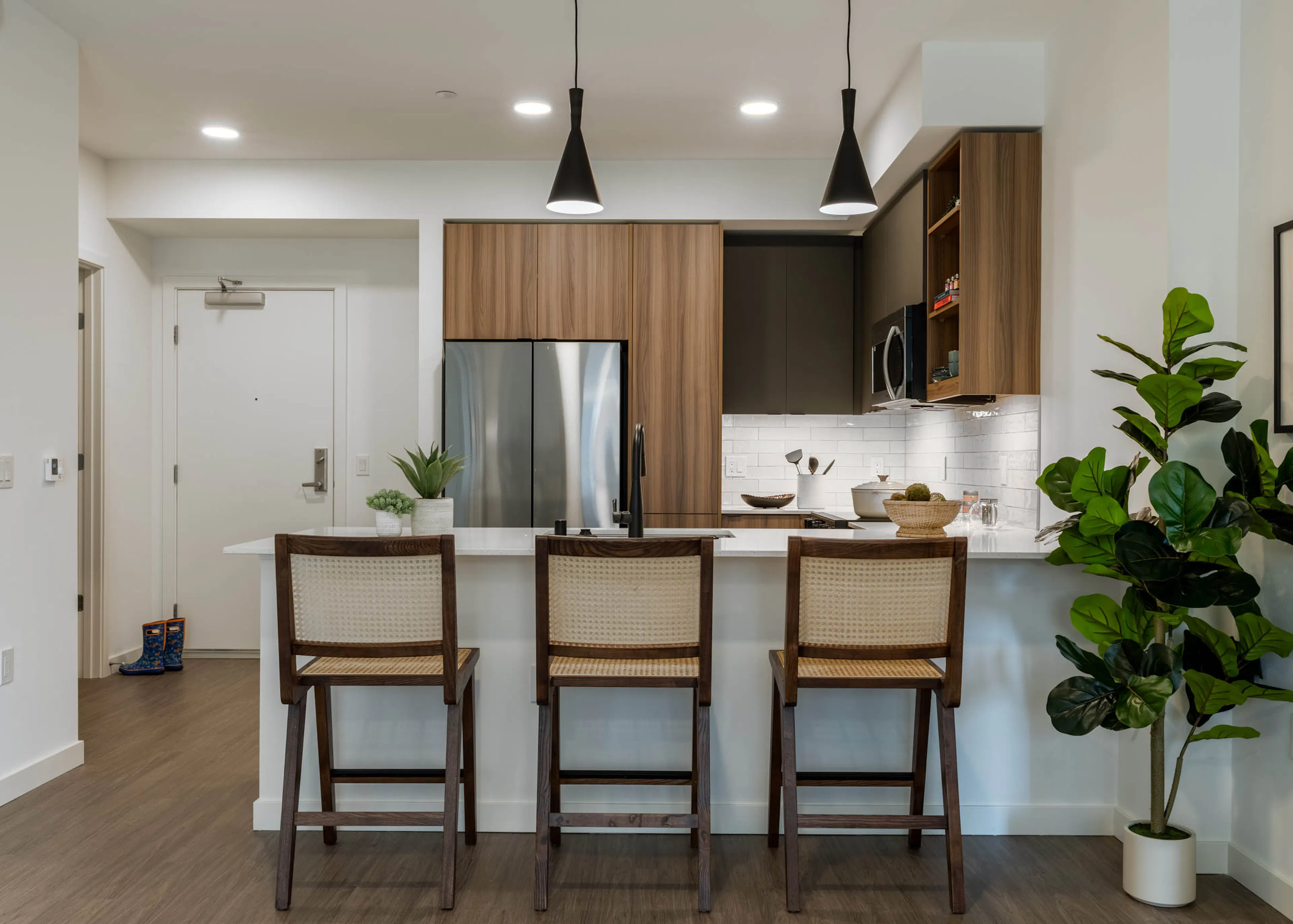 Kitchen with stainless steel appliances, wooden brown and grey cabinetry large white countertop and 3 highchairs and hanging spotlights above.