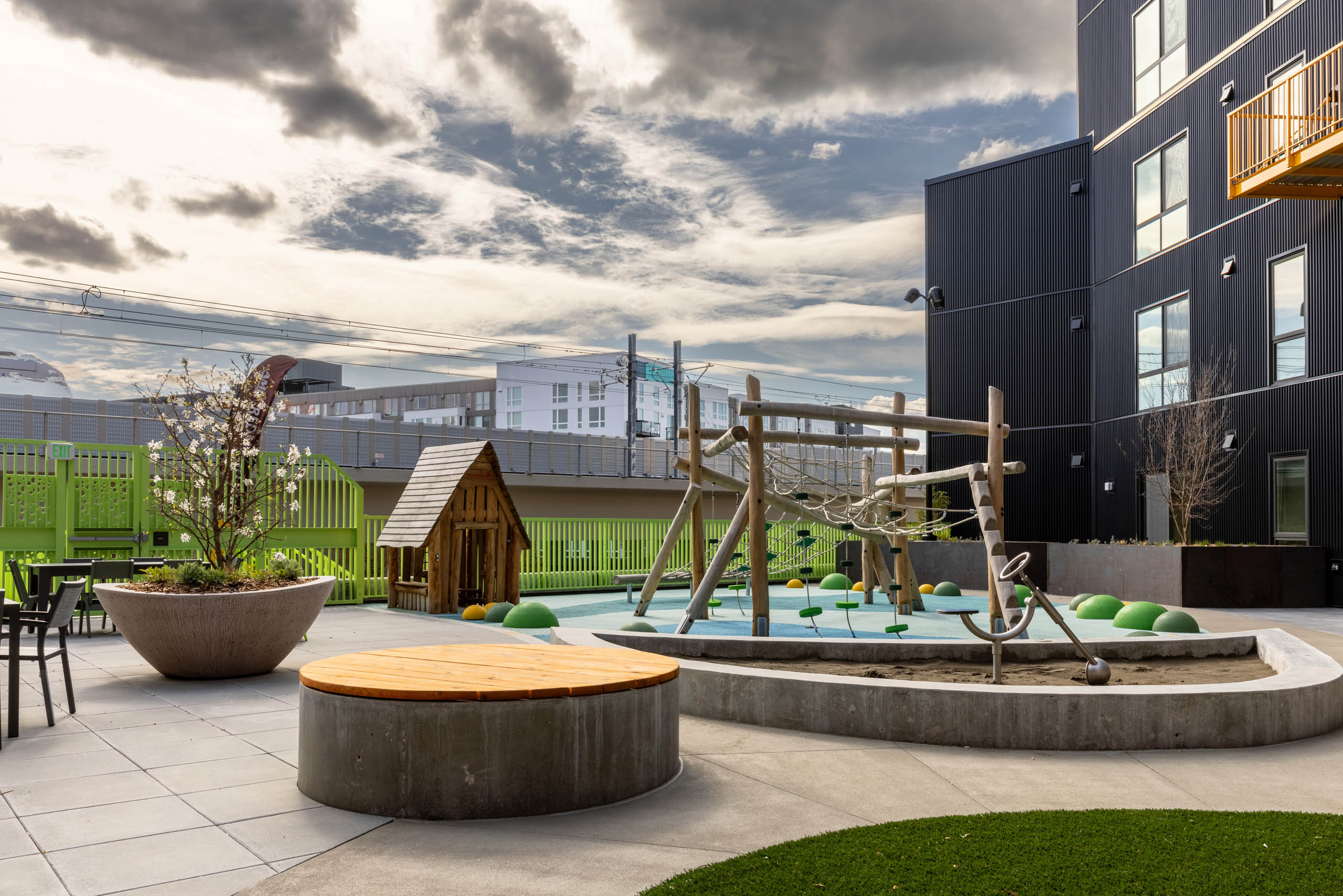 Outdoor play area with a climbing frame and a playhouse next to a green fence with seating available nearby.