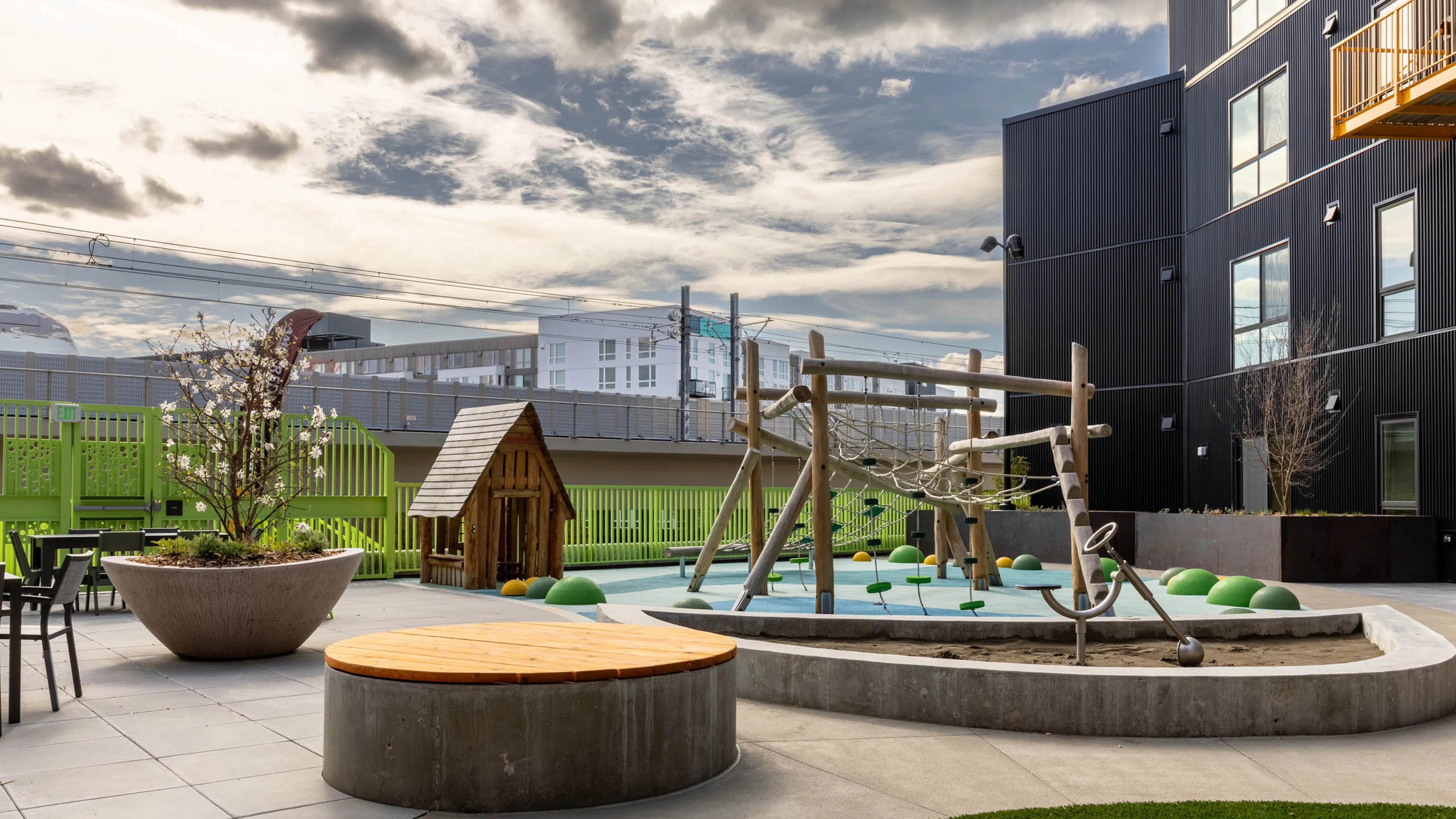 Outdoor play area with a climbing frame and a playhouse next to a green fence with seating available nearby.