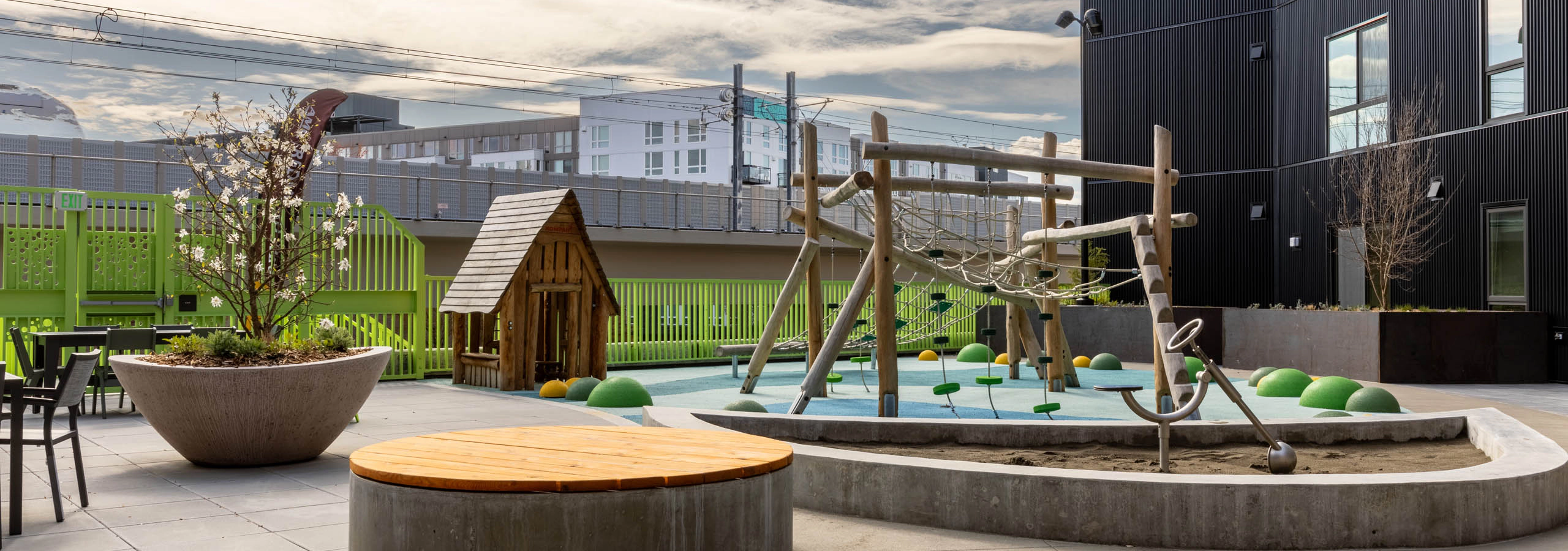 Outdoor play area with a climbing frame and a playhouse next to a green fence with seating available nearby.