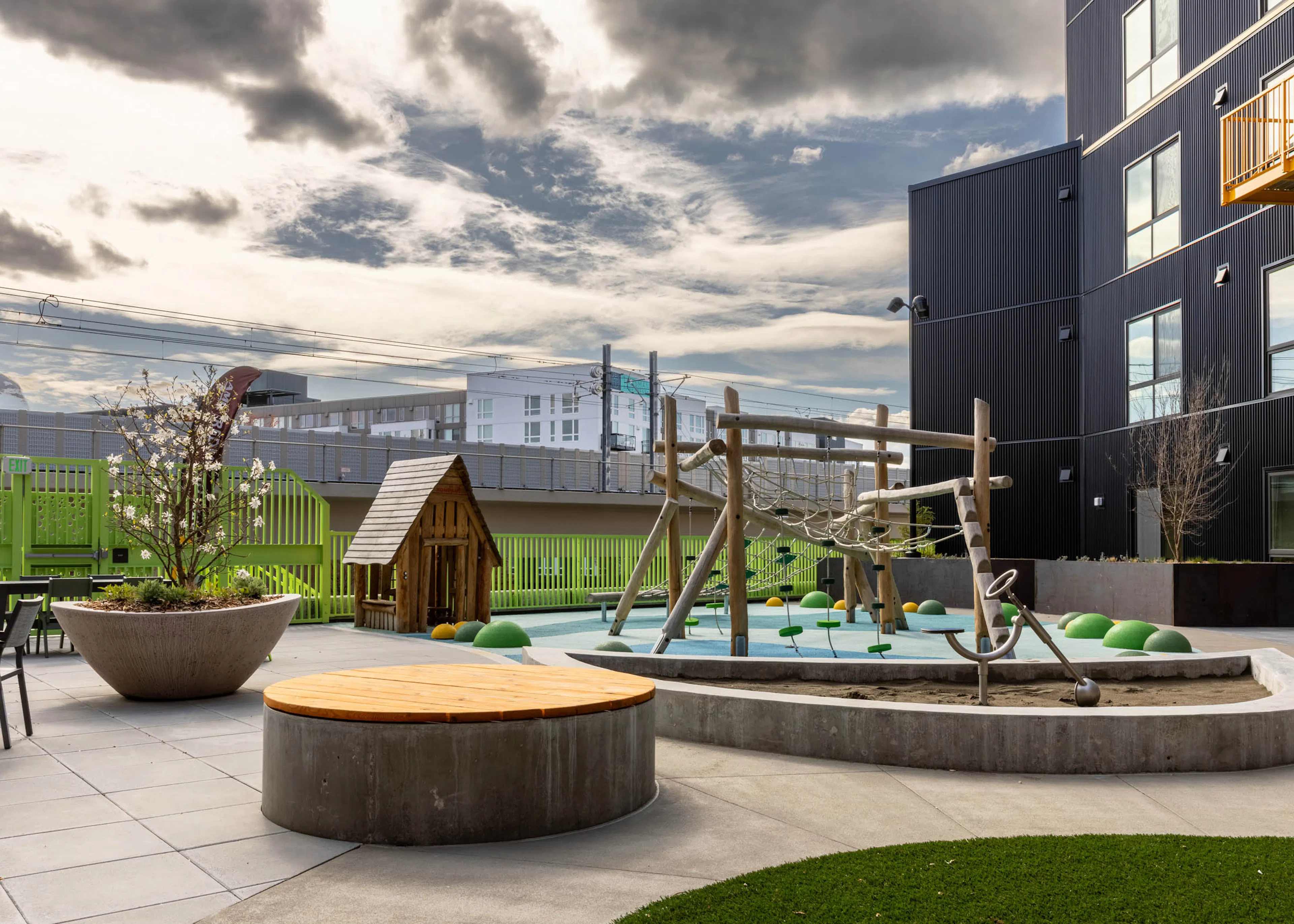 Outdoor play area with a climbing frame and a playhouse next to a green fence with seating available nearby.