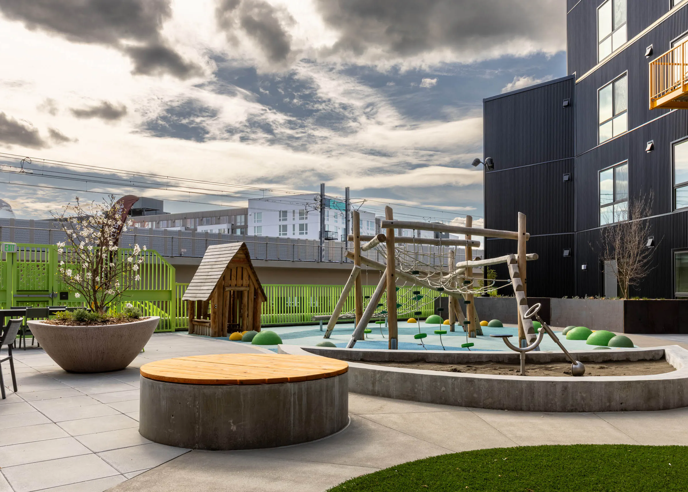 Outdoor play area with a climbing frame and a playhouse next to a green fence with seating available nearby.