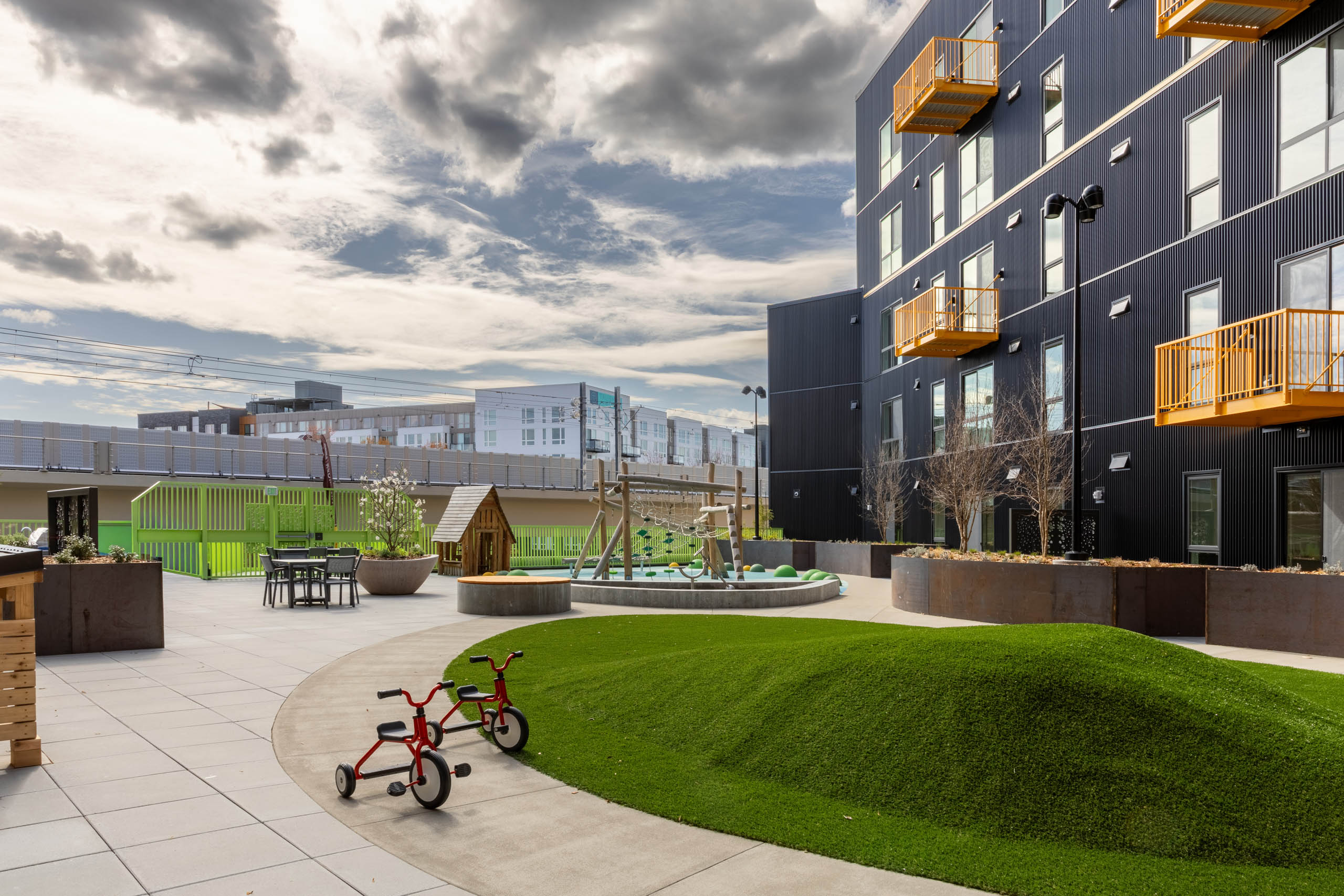 Outdoor play area with a turfed mound, climbing frame and a playhouse next to a green fence with seating available nearby.