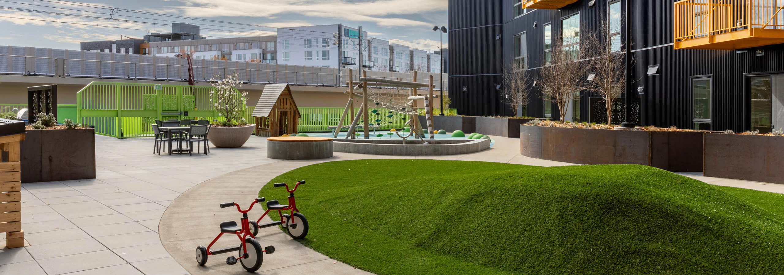 Outdoor play area with a turfed mound, climbing frame and a playhouse next to a green fence with seating available nearby.