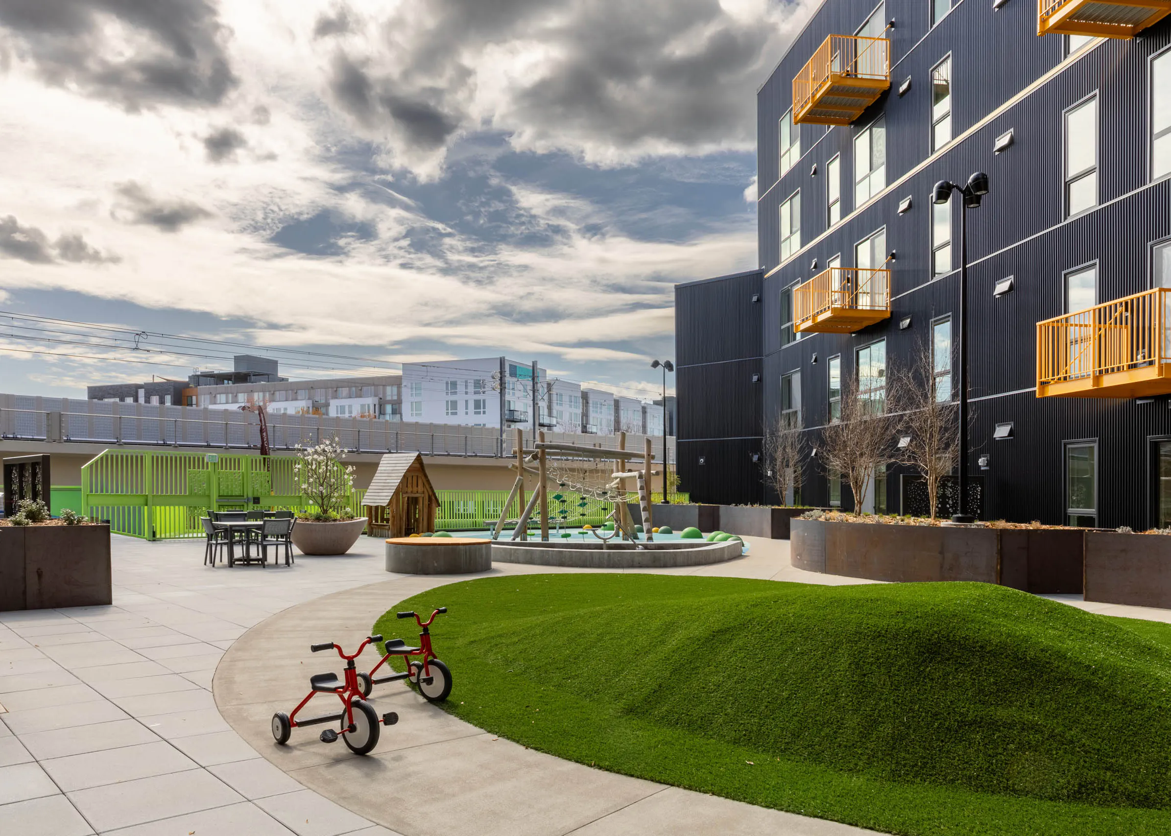 Outdoor play area with a turfed mound, climbing frame and a playhouse next to a green fence with seating available nearby.
